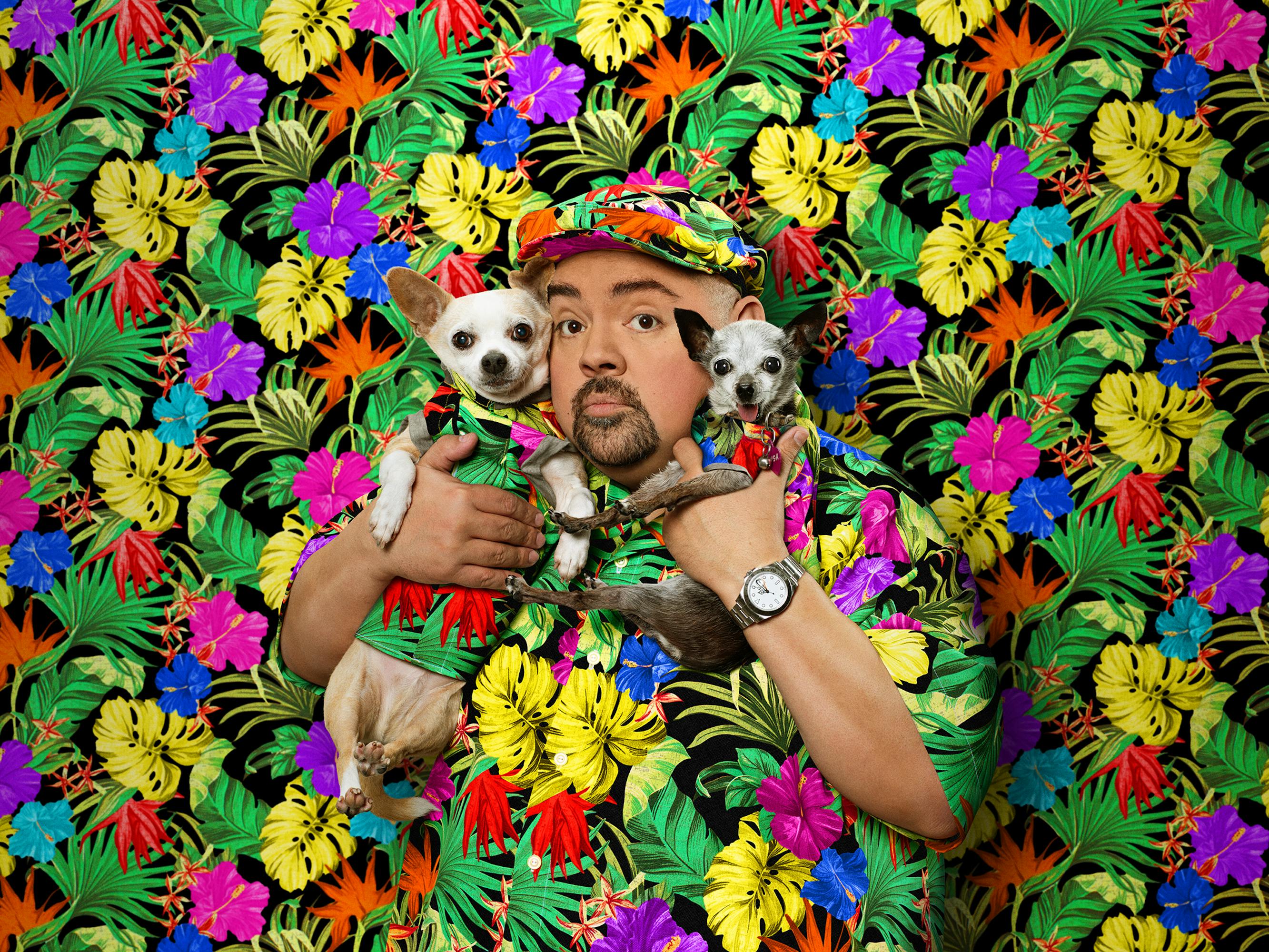 Gabriel Iglesias blends in with a wall of flowers, holding his dogs close to his face.