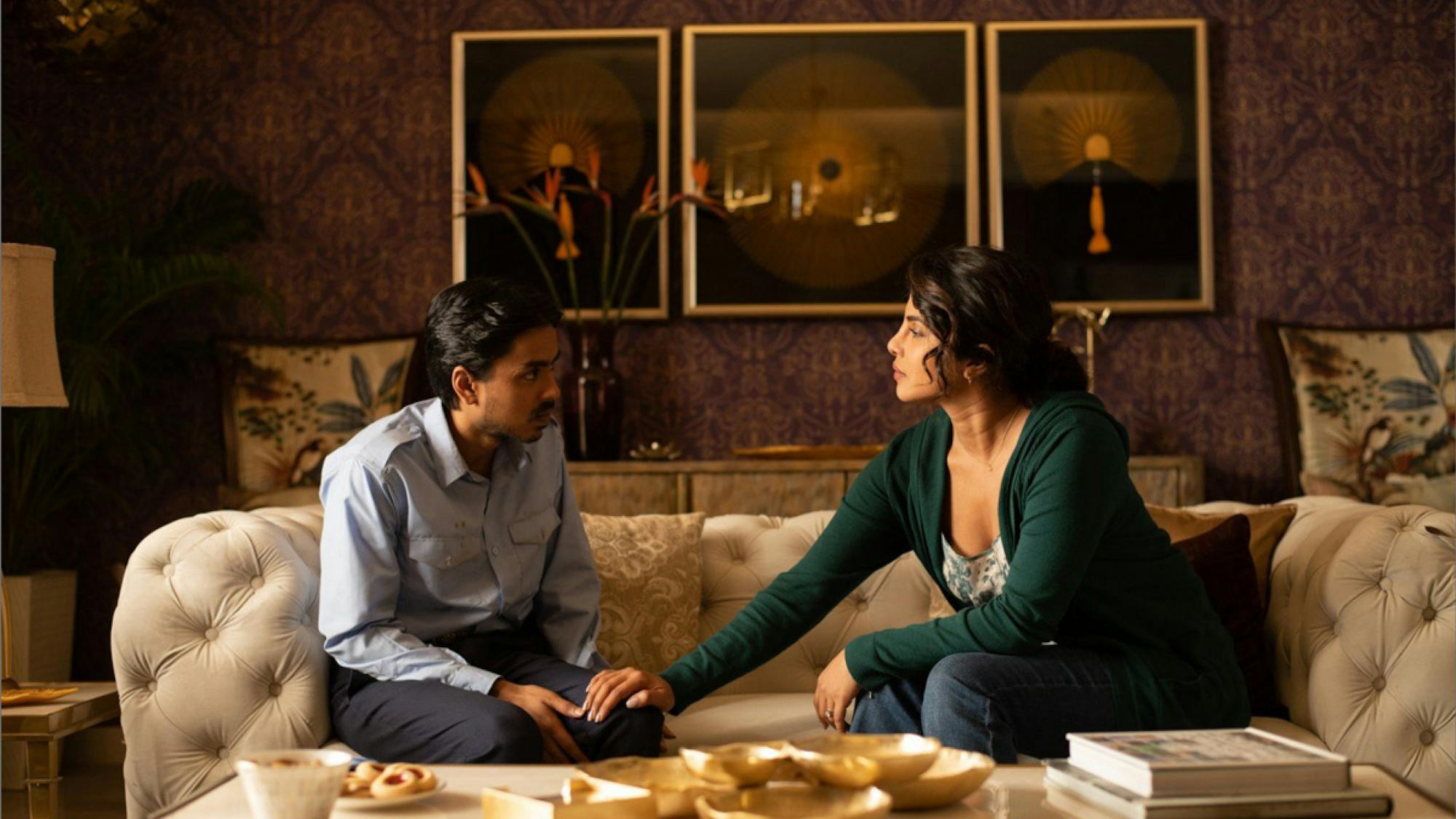 Priyanka Chopra Jonas, as Pinky Madam, is sitting on a couch with Balram, played by Adarsh Gourav. They are facing each other and she has her hand reached out on his knee as if she is comforting him. They are in a nicely decorated room and a coffee table in front of them looks to have some snacks set out.