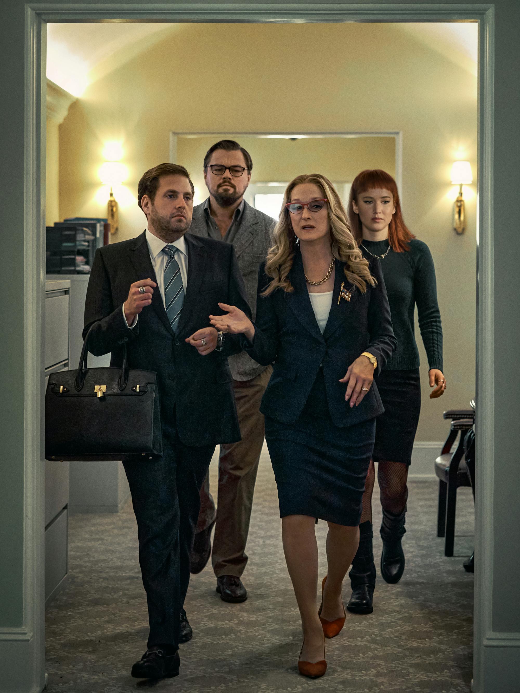 Jonah Hill, Leonardo DiCaprio, Meryl Streep, and Jennifer Lawrence walk through the Oval Office hallway. Hill wear a suit and carries a black Birkin. DiCaprio wears a grey blazer and khakis. Streep wears a navy skirt suit. Lawrence wears a grey turtleneck and black shirt. 