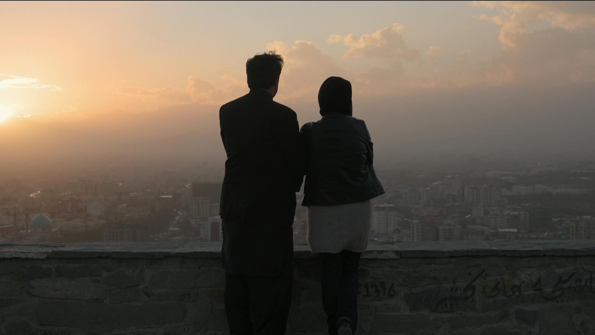 Two people stands on a roof looking out on the city, as the sun rises or sets in the distance.