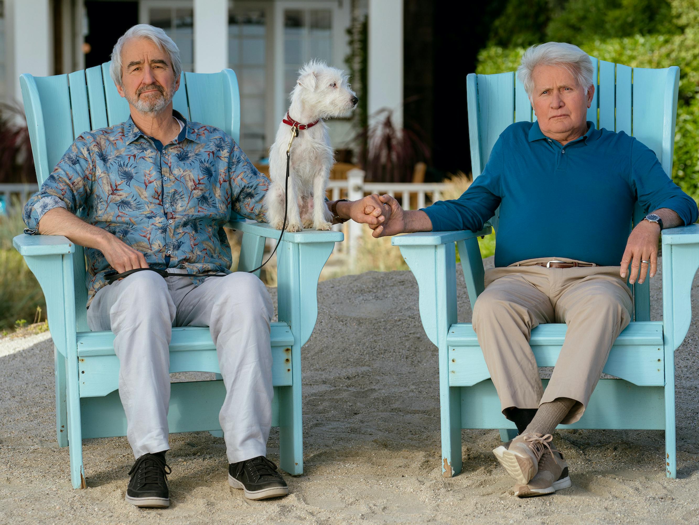 Sam Waterston and Martin Sheen sit in light blue chairs holding hands. A white dog perches on the arm of Sam's chair. Sam wears grey pants, black shoes, and a patterned shirt. Martin wears khakis and a teal shirt. They are the picture of marital bliss in the beachy shot. 