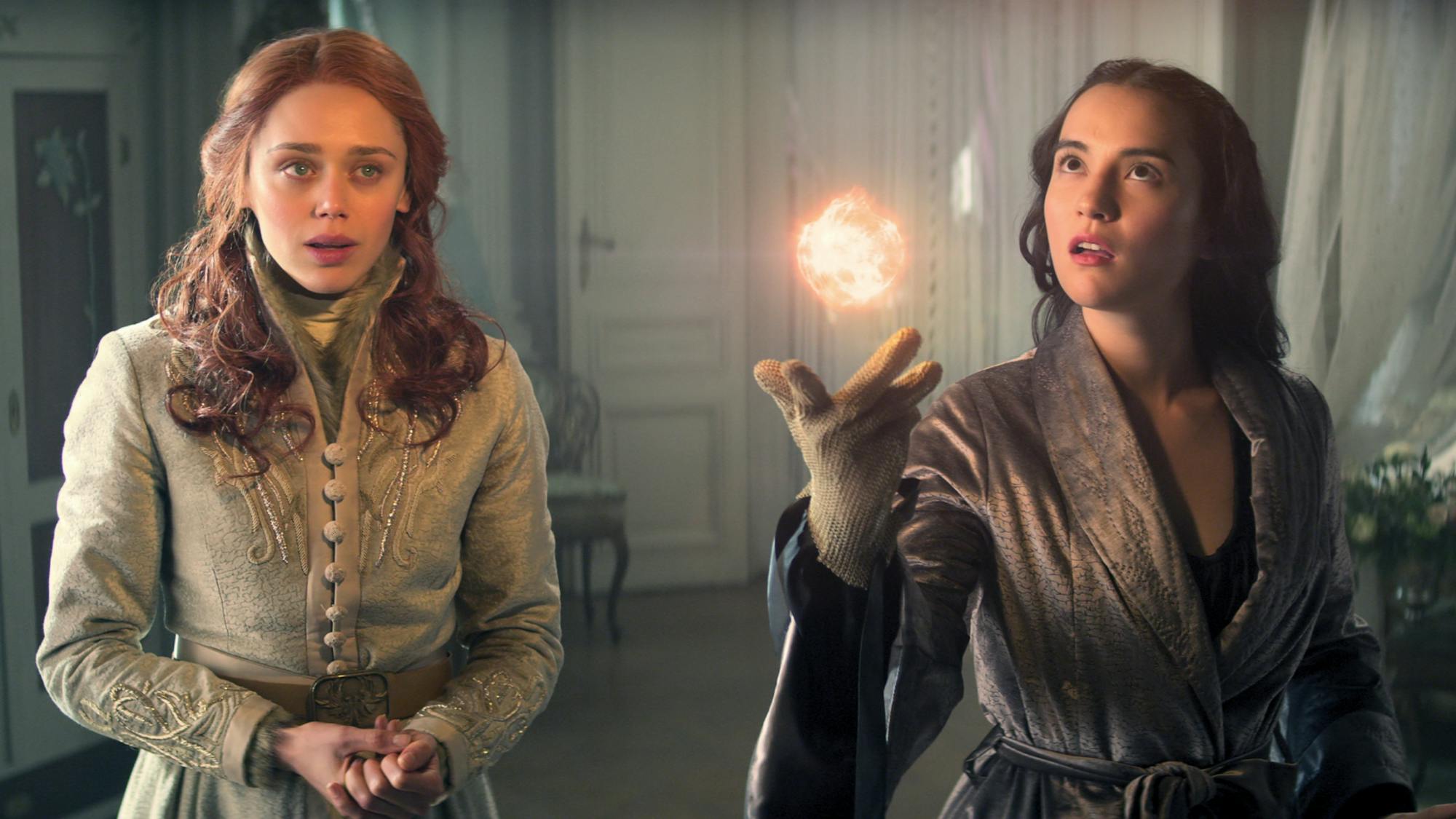 Daisy Head playing Genya Safin and Jessie Mei Li playing Alina Starkov stand side by side. They wear a light dress and dark robe respectively, and Mei Li hovers an orb of light above her outstretched hand. Both characters look surprised.