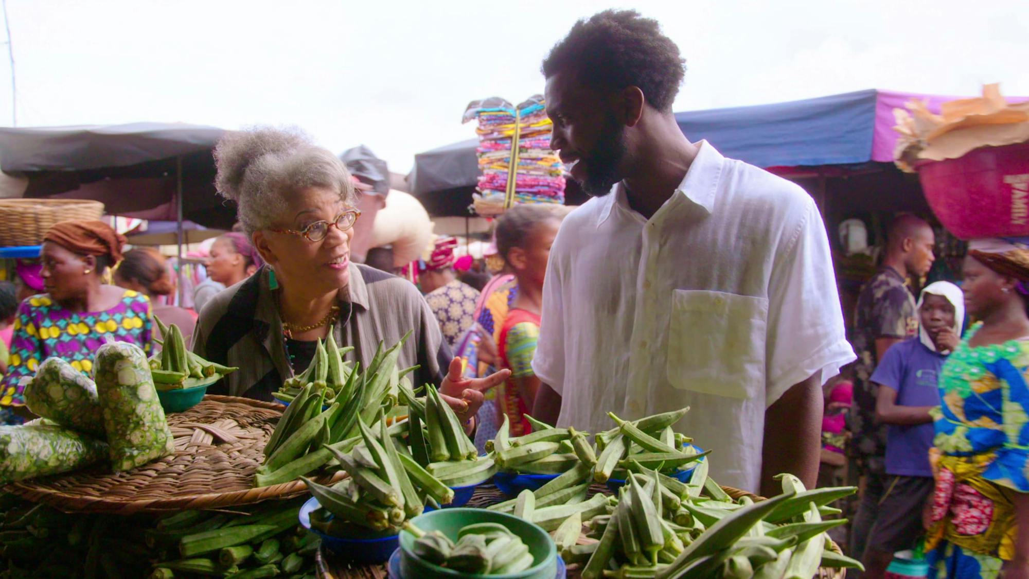 Jessica B. Harris and Stephen Satterfield shop in an outdoor market. They look at each other in conversation over a heaping pile of okra. In the background are lots of people dressed in bright colors.