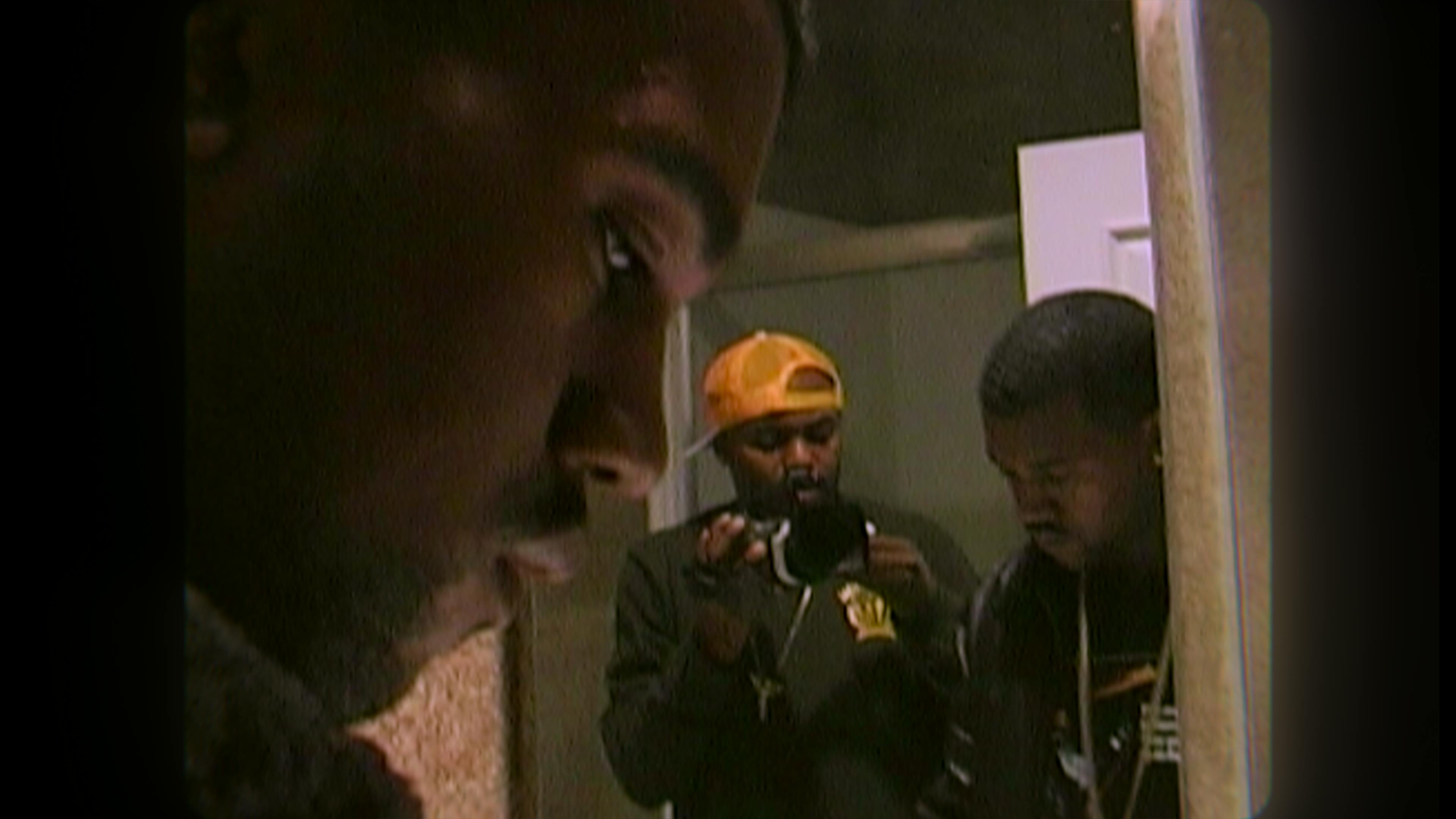 Kanye West looks down in a dark top. He's filmed by Coodie who wears a yellow hat.