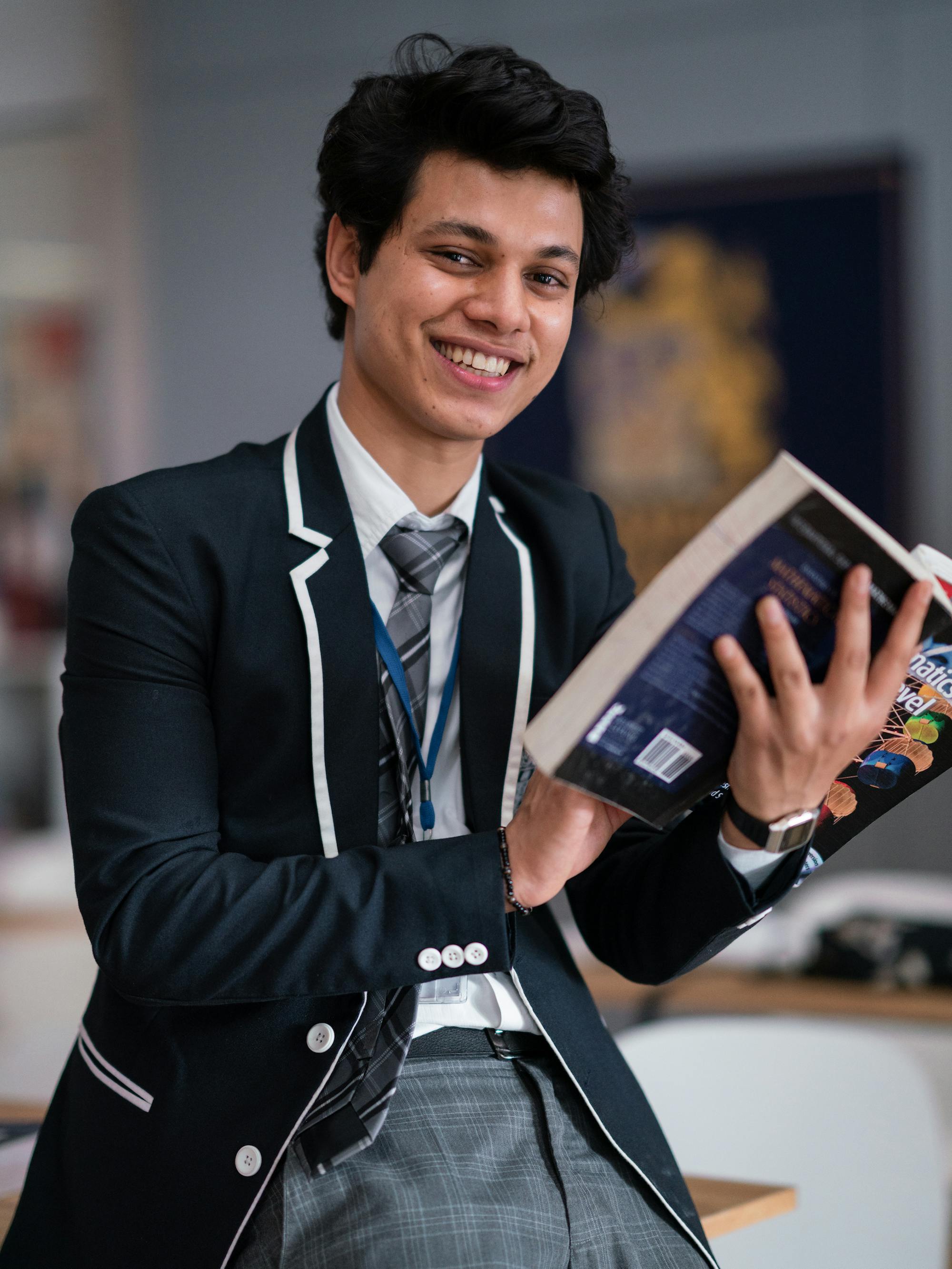 Actor Piyush Khati leans against a desk in a navy and gray school uniform, holding a book and smiling at the camera. 