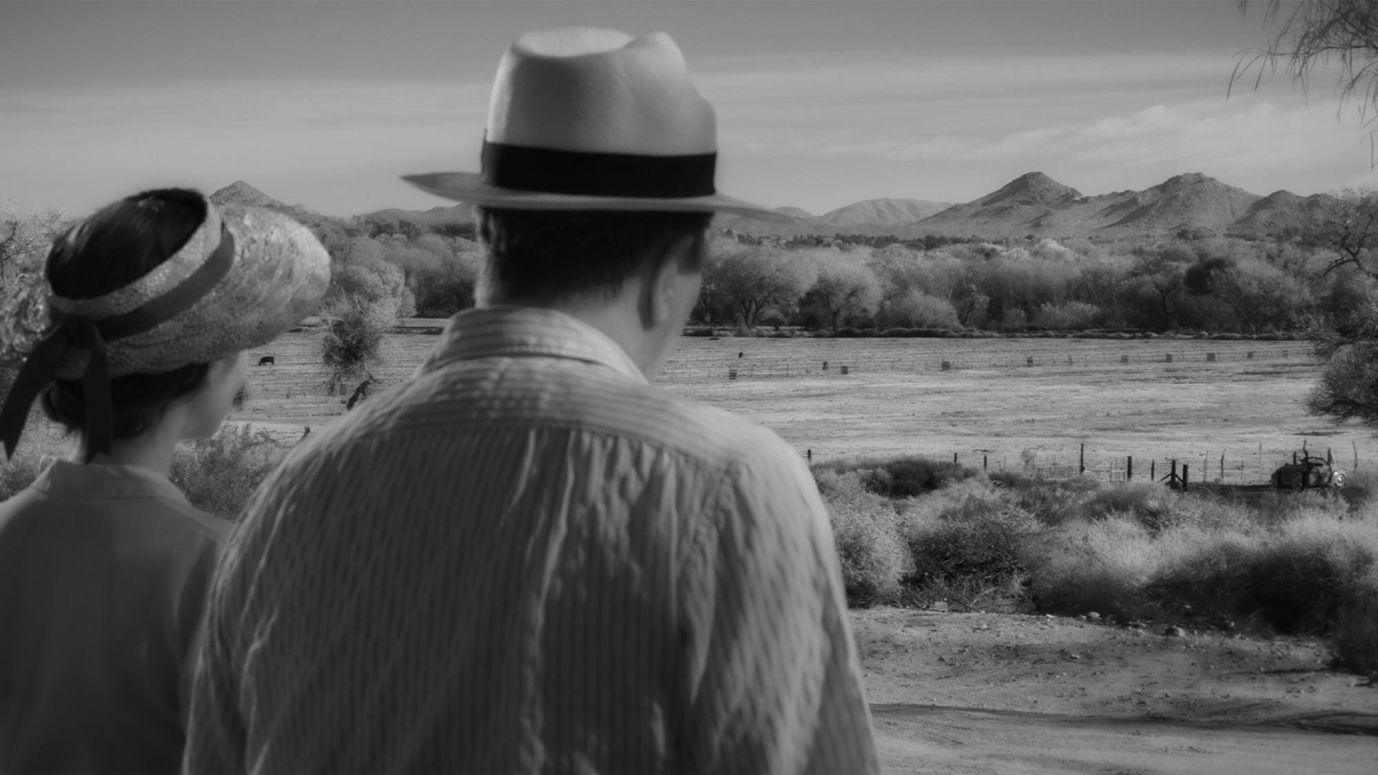 Rita and Mank look out at the landscape at Victorville. In the distance there are mountains and in the foreground there is a flat expanse of land. We see them from behind, both wearing hats.