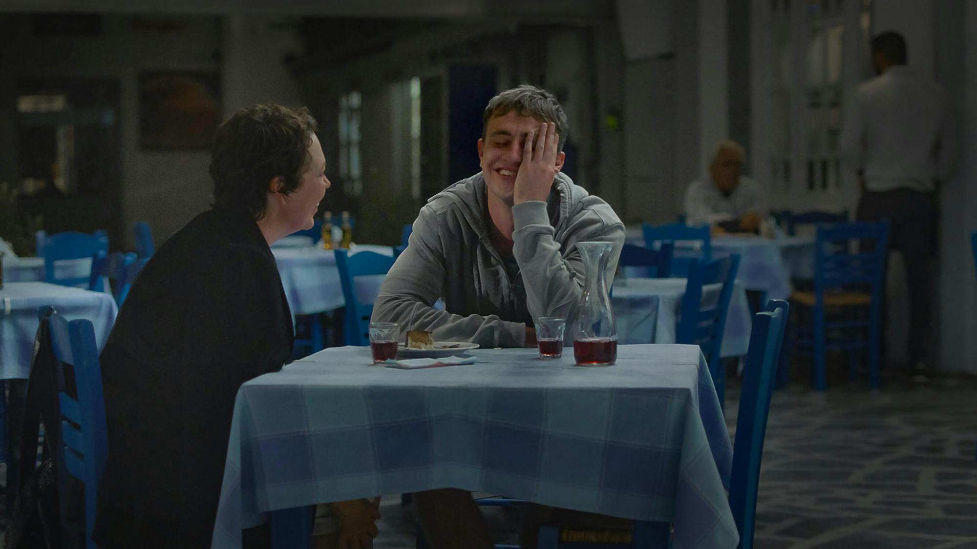 Leda (Olivia Colman) and Will (Paul Mescal) sit at a blue-checkered table-clothed table drinking red wine from a carafe and laughing.