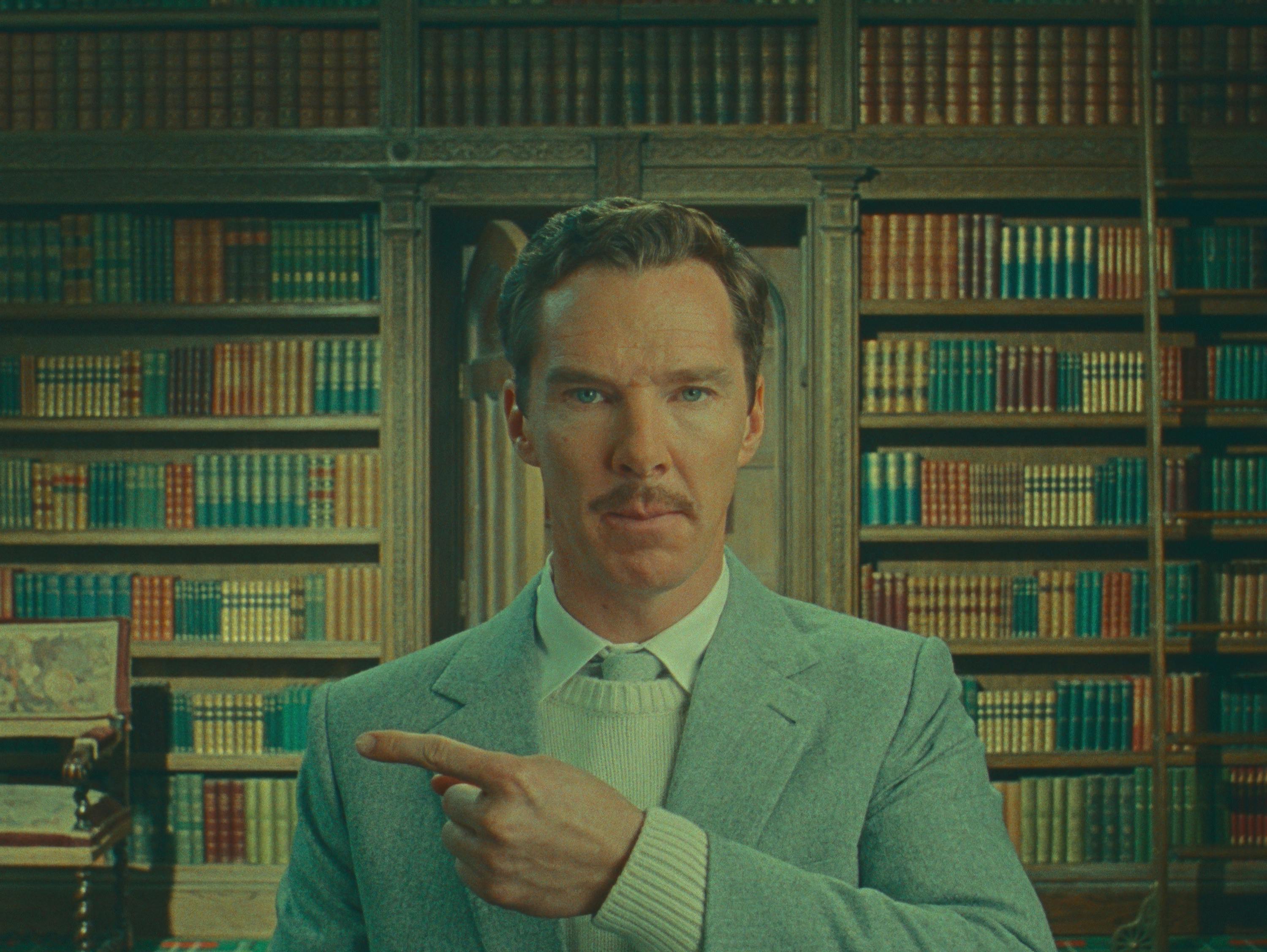 Henry Sugar (Benedict Cumberbatch) points at something offscreen and wears a sweater, blazer, tie combo.