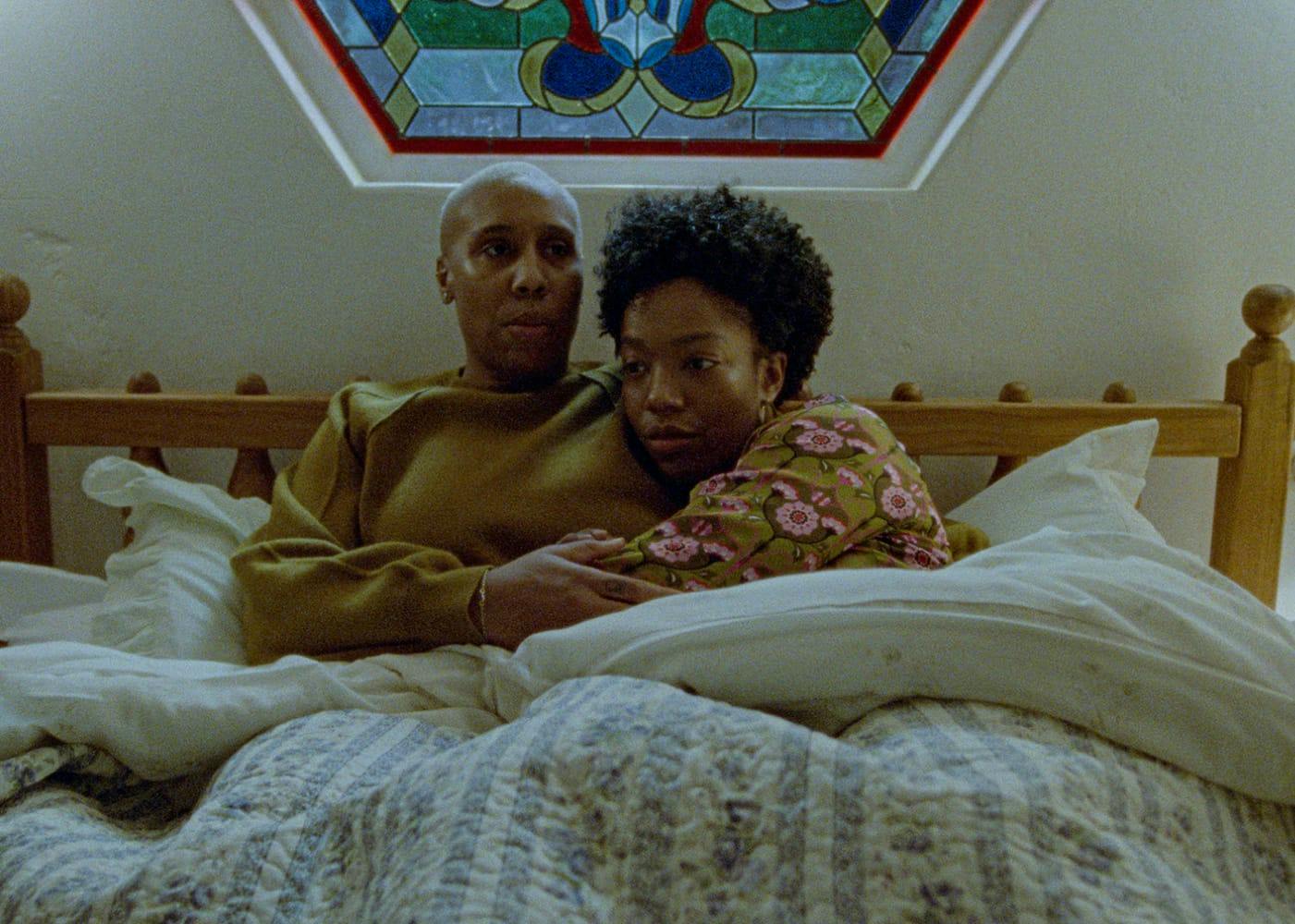 Denise (Lena Waithe) and Alicia (Naomi Ackie) lie in bed together. Alicia hugs Denise’s middle. Alicia wears a patterened pajama top, and Denise wears a yellow sweatshirt. Their bed is made of wood and they have white bedding.