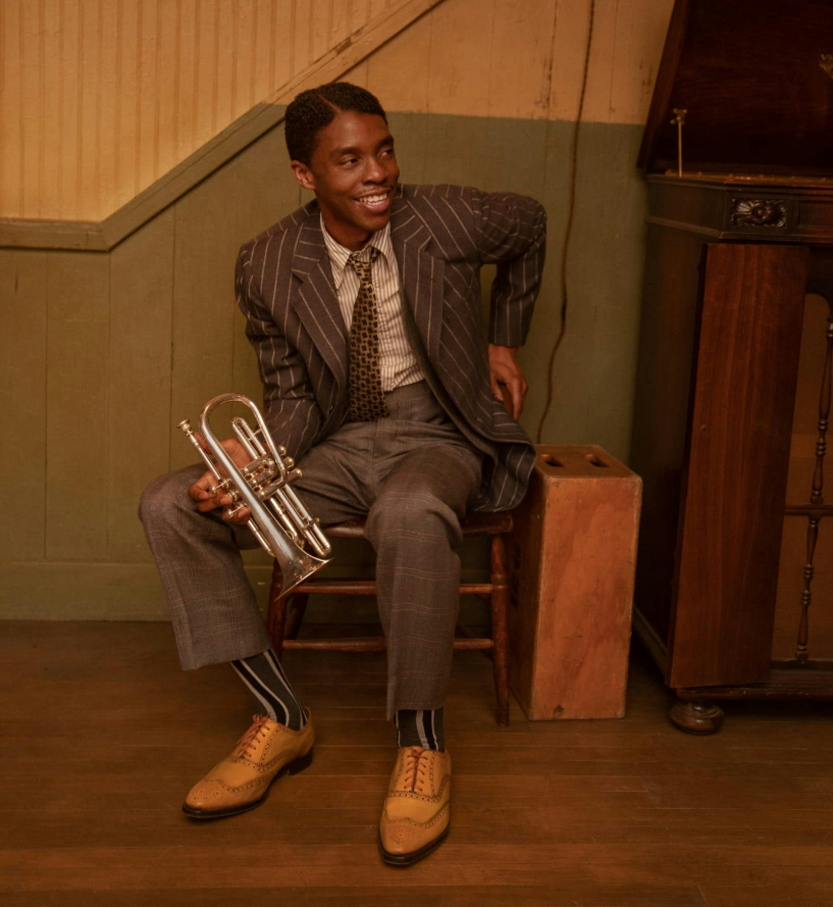 Chadwick Boseman as Levee in Ma Rainey’s Black Bottom. He wears a striped blazer, checkered pants, tan shoes, striped socks, and holds a trumpet. He sits in a wooden chair and smiles at someone off camera. 