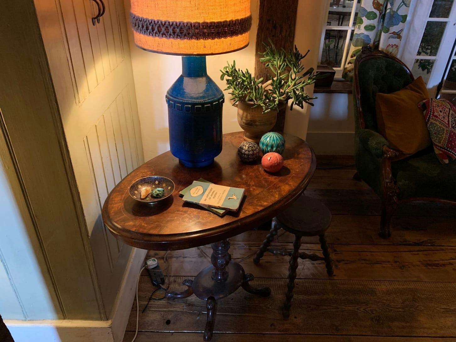 A wooden table with a blue lamp, plant, three decorative balls, some books, and a dish. There is a stool to the side, and a green chair against the window.