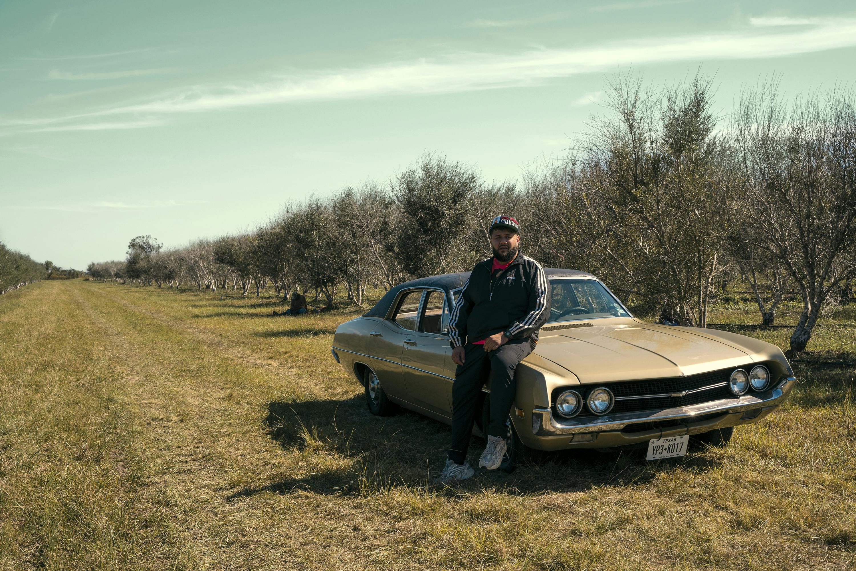 Mo Amer sits against his brown classic car on the olive farm. The sky is light blue streaked with clouds. Amer wears a black Adidas tracksuit and a hat.