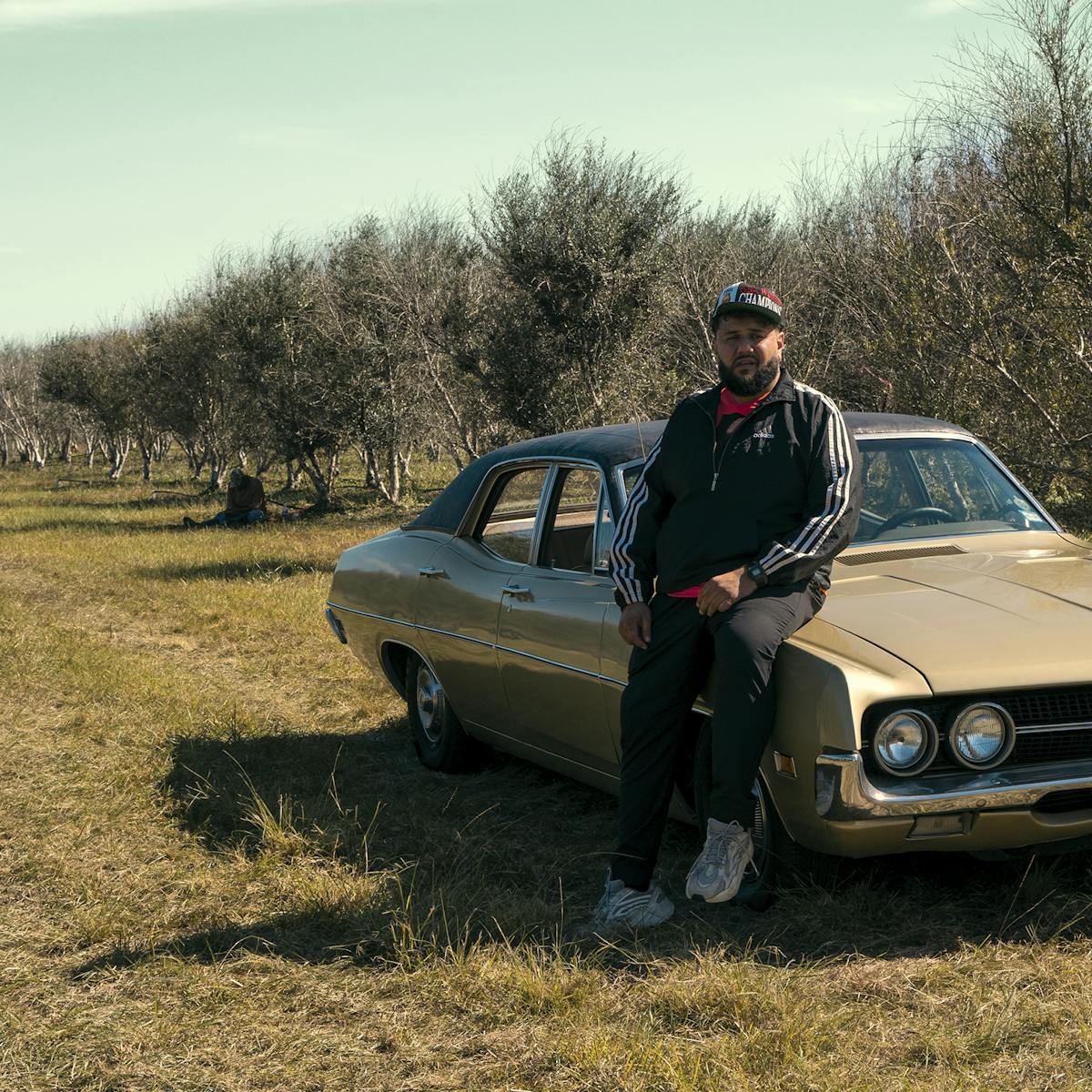 Mo Amer sits against his brown classic car on the olive farm. The sky is light blue streaked with clouds. Amer wears a black Adidas tracksuit and a hat.