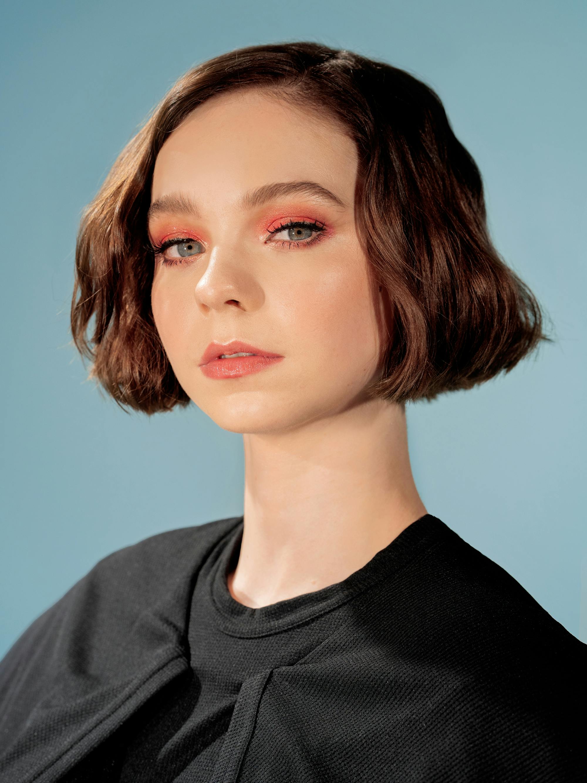 Emma Myers wears a black top and red eye shadow, against a blue background.