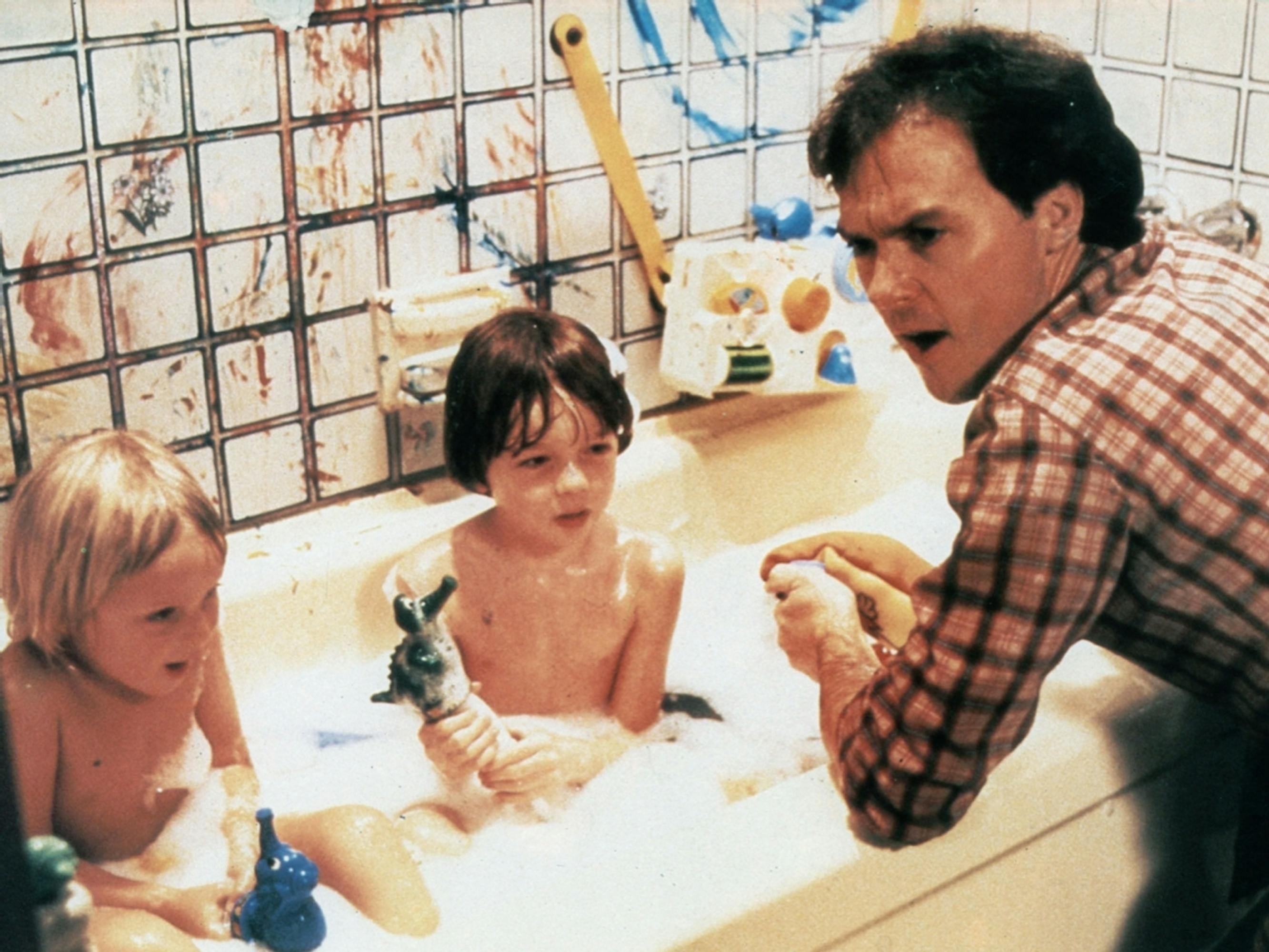 Jack Butler (Michael Keaton) in Mr. Mom. Butler wears a checkered shirt as he sits besides his childrens bath.
