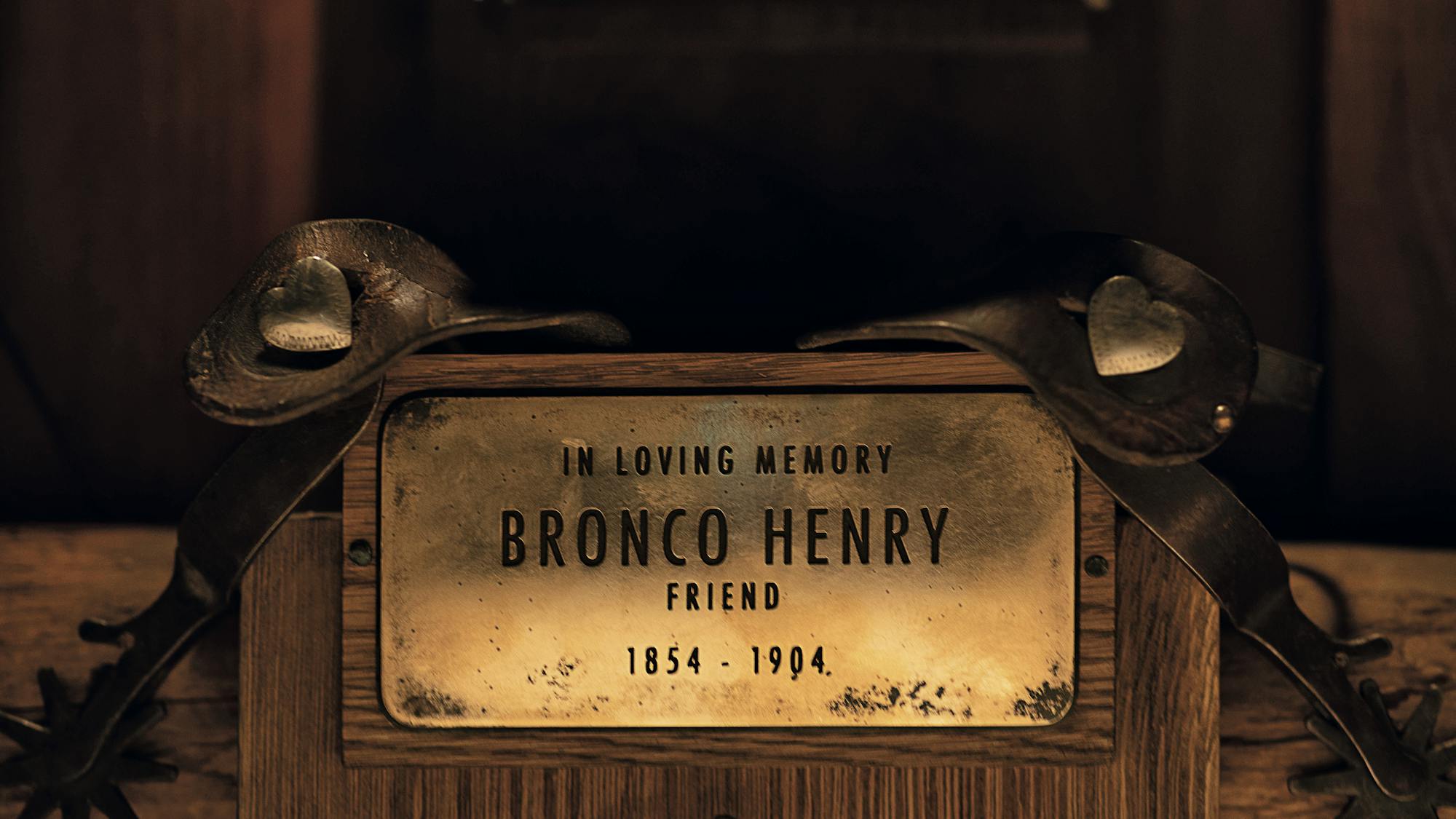 A gold Bronco Henry nameplate on a wooden saddle. There are leather straps around the nameplate with little heart shaped studs. 