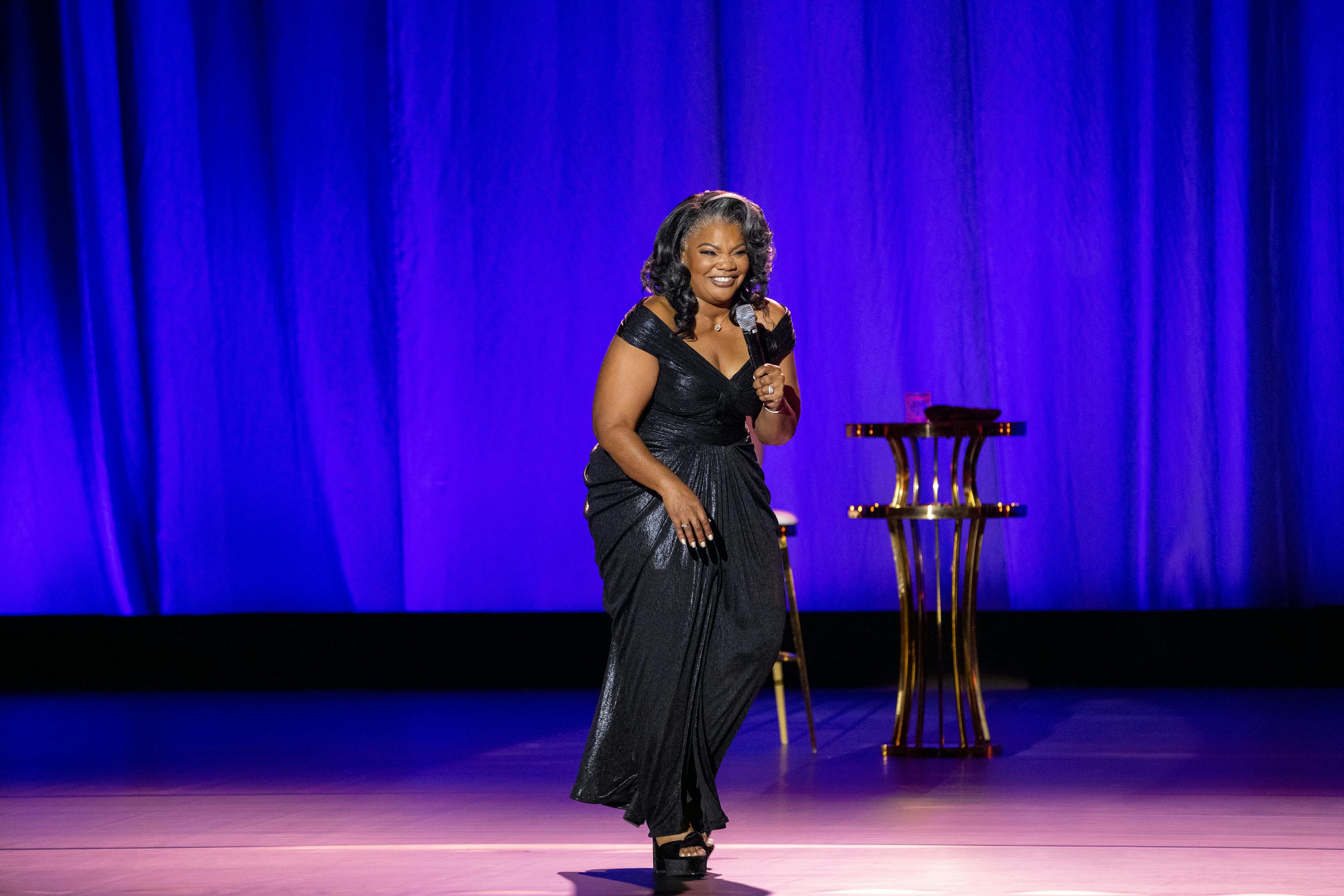 Mo’Nique wears a long black dress and black heels as she moves across a stage laughing into a mic.