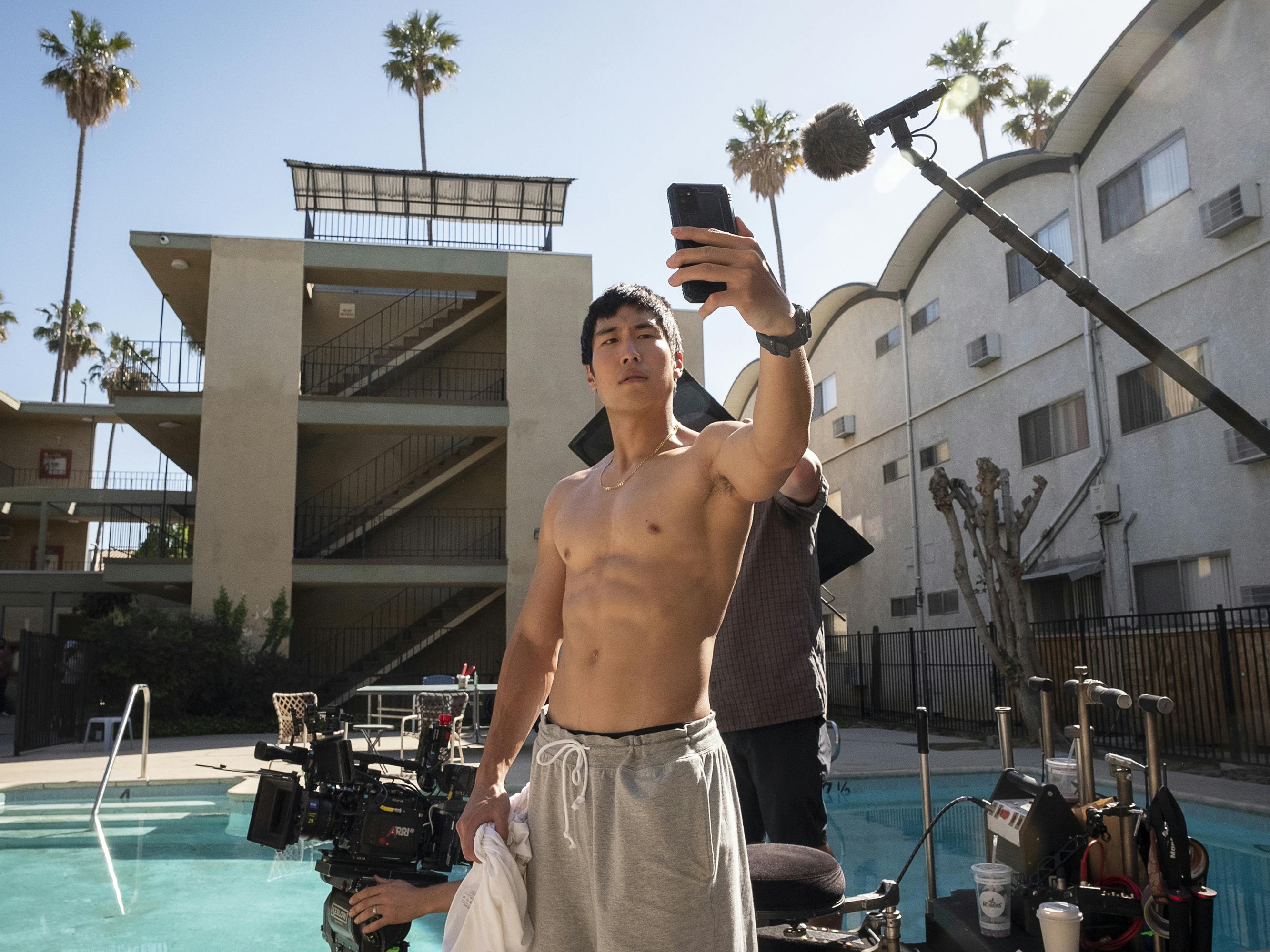 Young Mazino takes a selfie poolside in this behind-the-scenes shot. The sun shines from behind his character’s beige apartment building, illuminating his muscles and rows of abs nicely. 