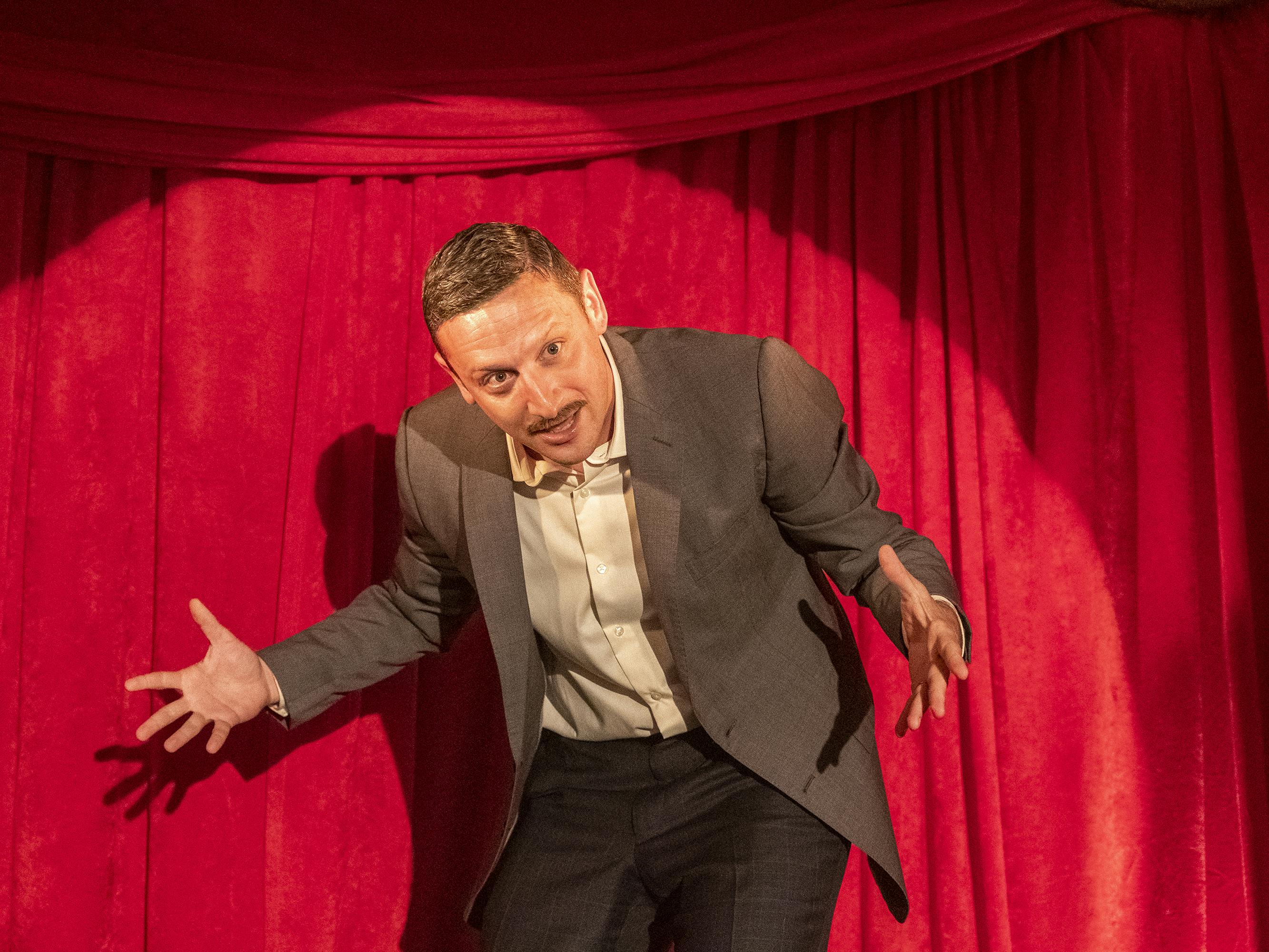 Tim Robinson stands in the spotlight against a red velvet curtain.