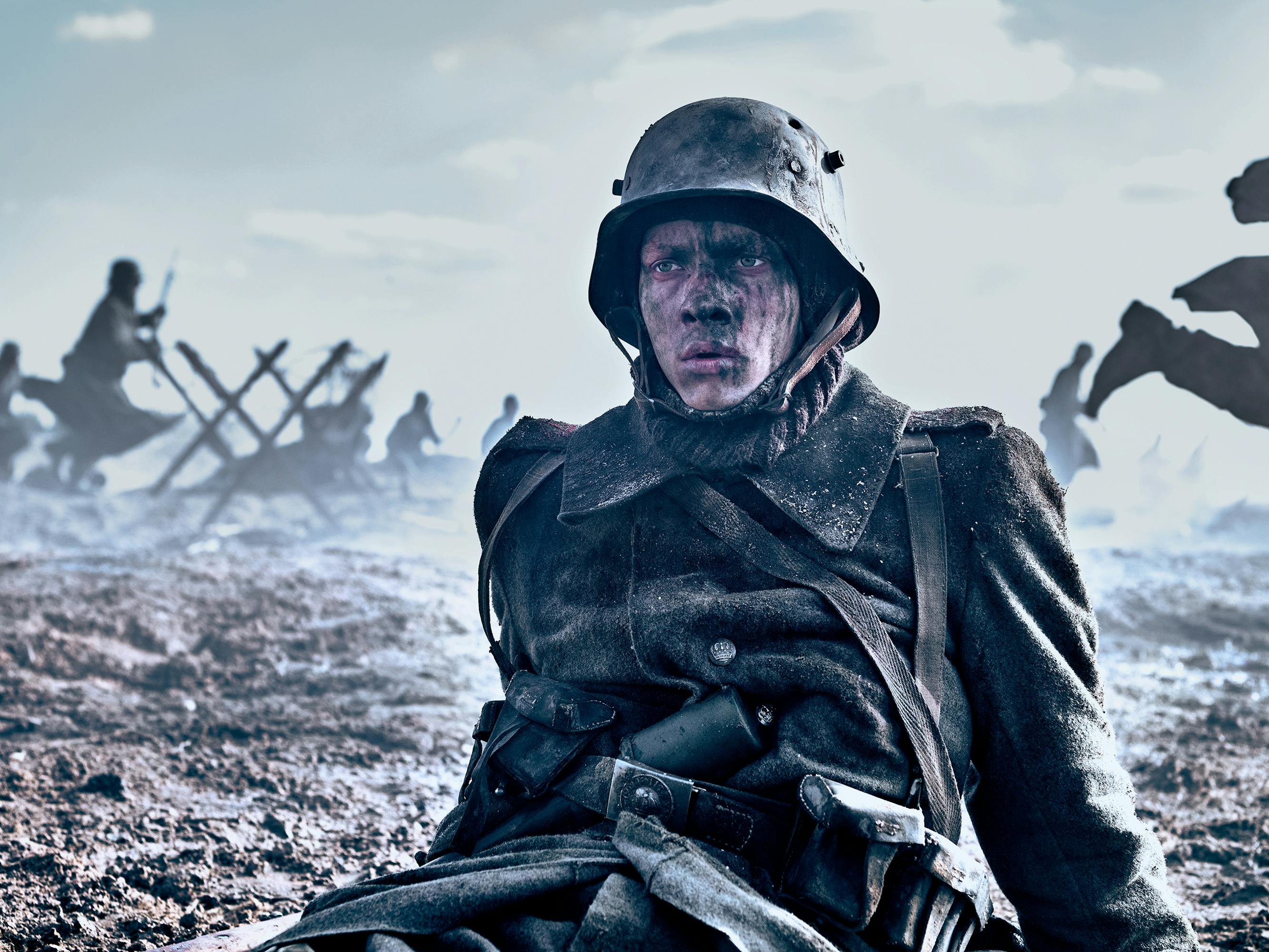 Paul Bäumer (Felix Kammerer) sits on the muddy ground in his green uniform and helmet. His face is streaked with mud, and behind him are soldiers running around and fighting.