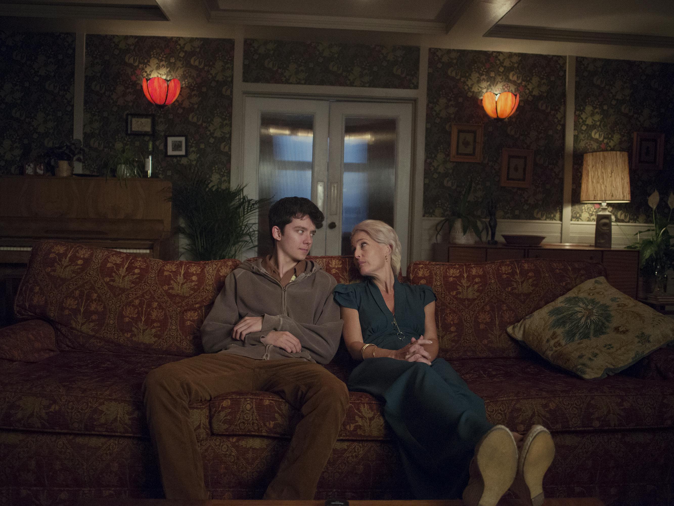 Otis Milburn (Asa Butterfield) and Dr. Jean Milburn (Gillian Anderson) sit side by side on a patterned couch in their dimly lit living room.