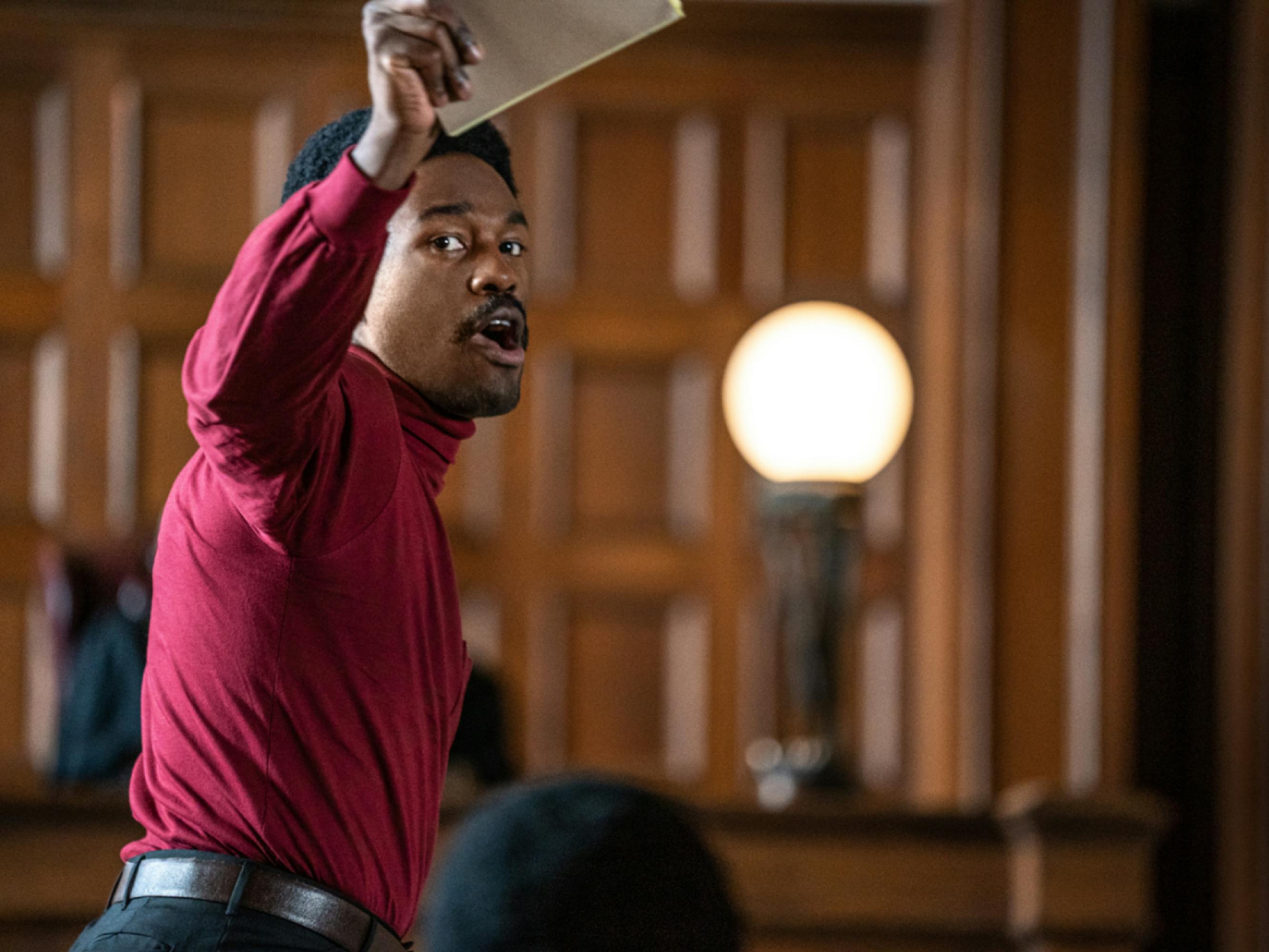 Abdul-Mateen as Bobby Seale in a courtroom scene from The Trial of the Chicago 7. He wears a red turtleneck and holds up a pad of paper, portraying Seale as he made the case that he was being denied his constitutional right to a defense.