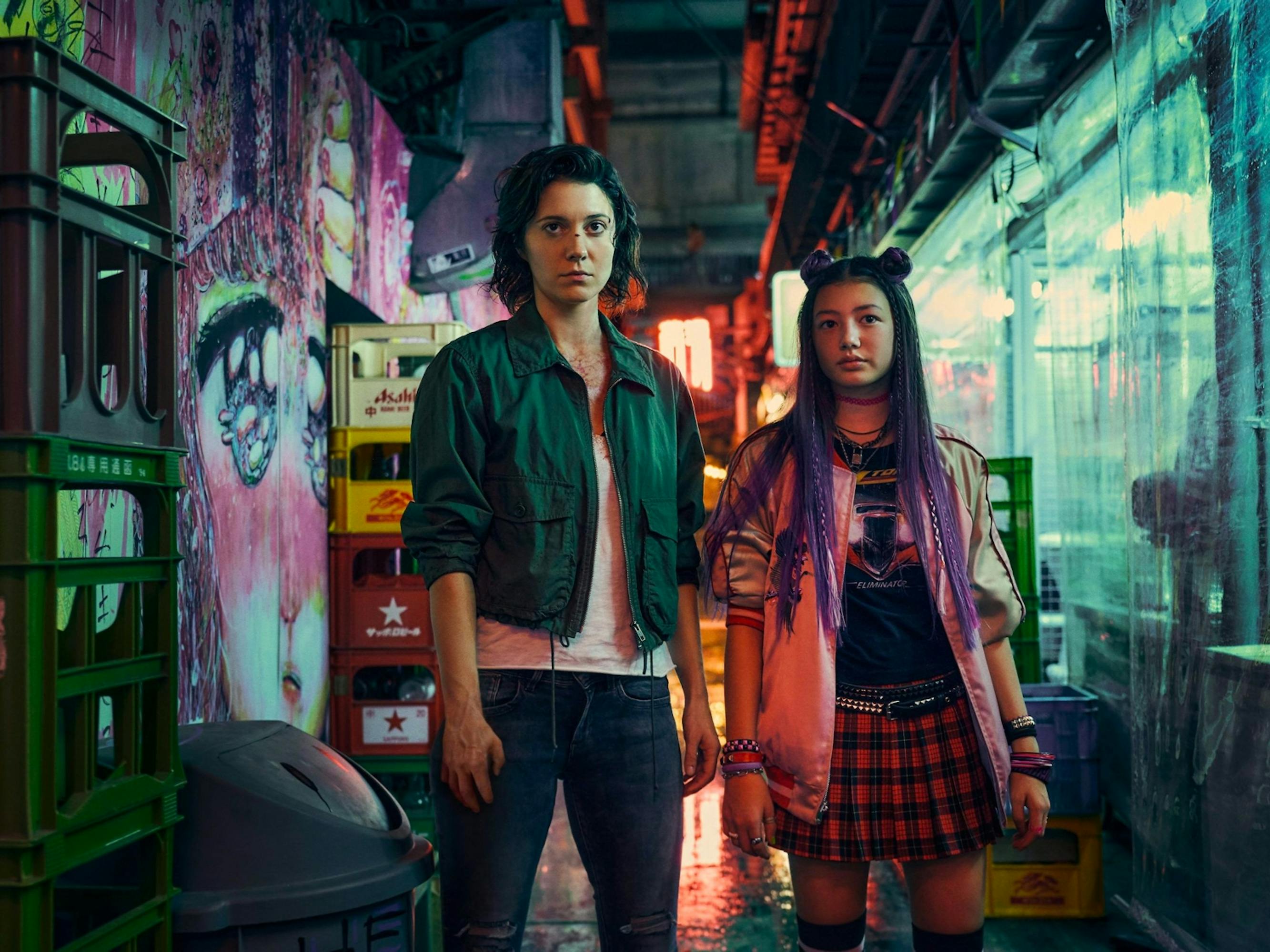 Kate (Mary Elizabeth Winstead) and Ani (Miku Martineau) stand in an alley crowded with stacked crates. On the left wall is a huge anime face, and on the right is hanging plastic. Kate wears jeans and a green jacket, and Ani wears a plaid skirt, pink jacket, and has purple hair.
