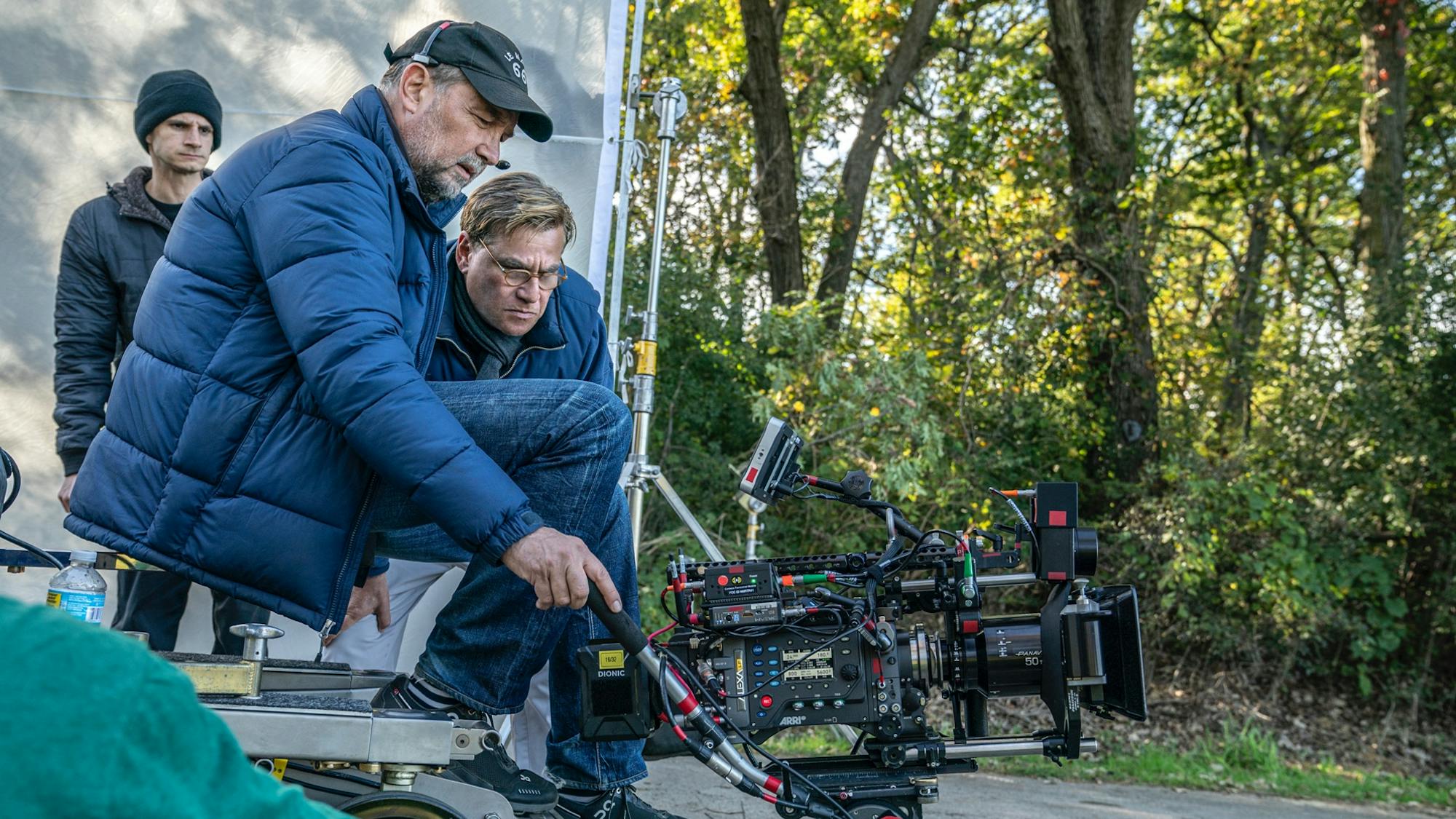 Papamichael is perched on a camera rig wearing blue jeans and a blue down jacket. Sorkin, also in blue, leans over to review a shot. Light shines through some trees behind them.