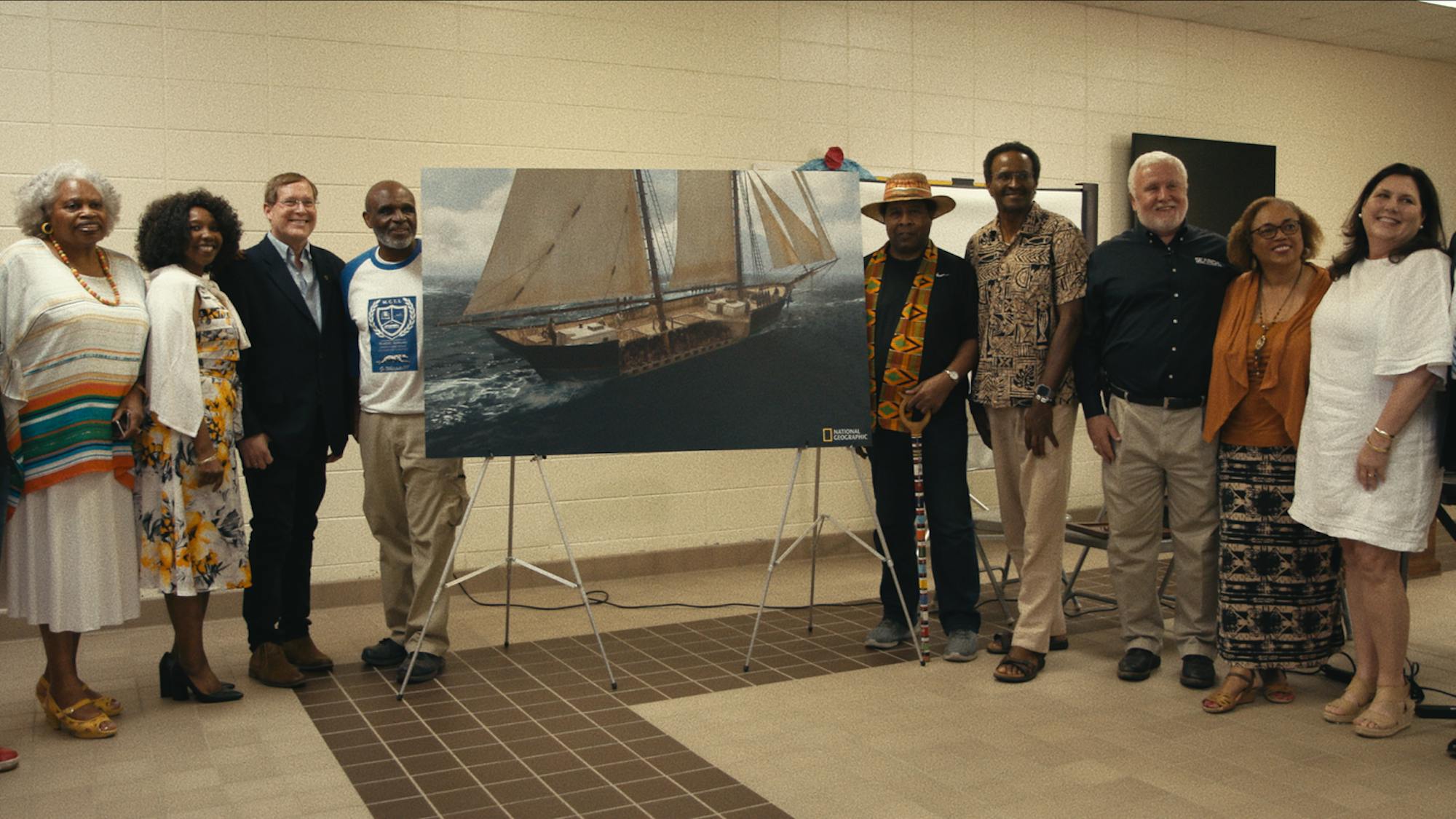 Some community members pose around a rendering of the Clotilda. They stand in a beige room facing the camera.