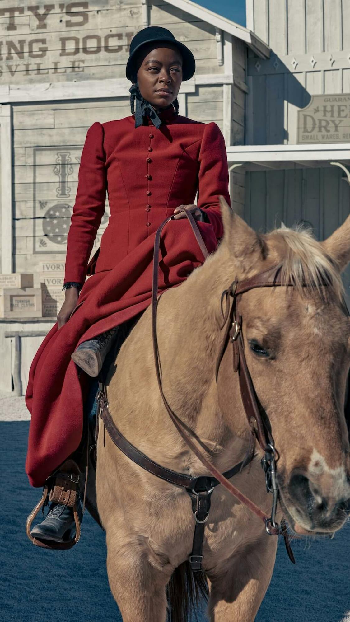 Cuffee (Danielle Deadwyler) wears a long red dress with little buttons and a black hat. She rides a brown horse.
