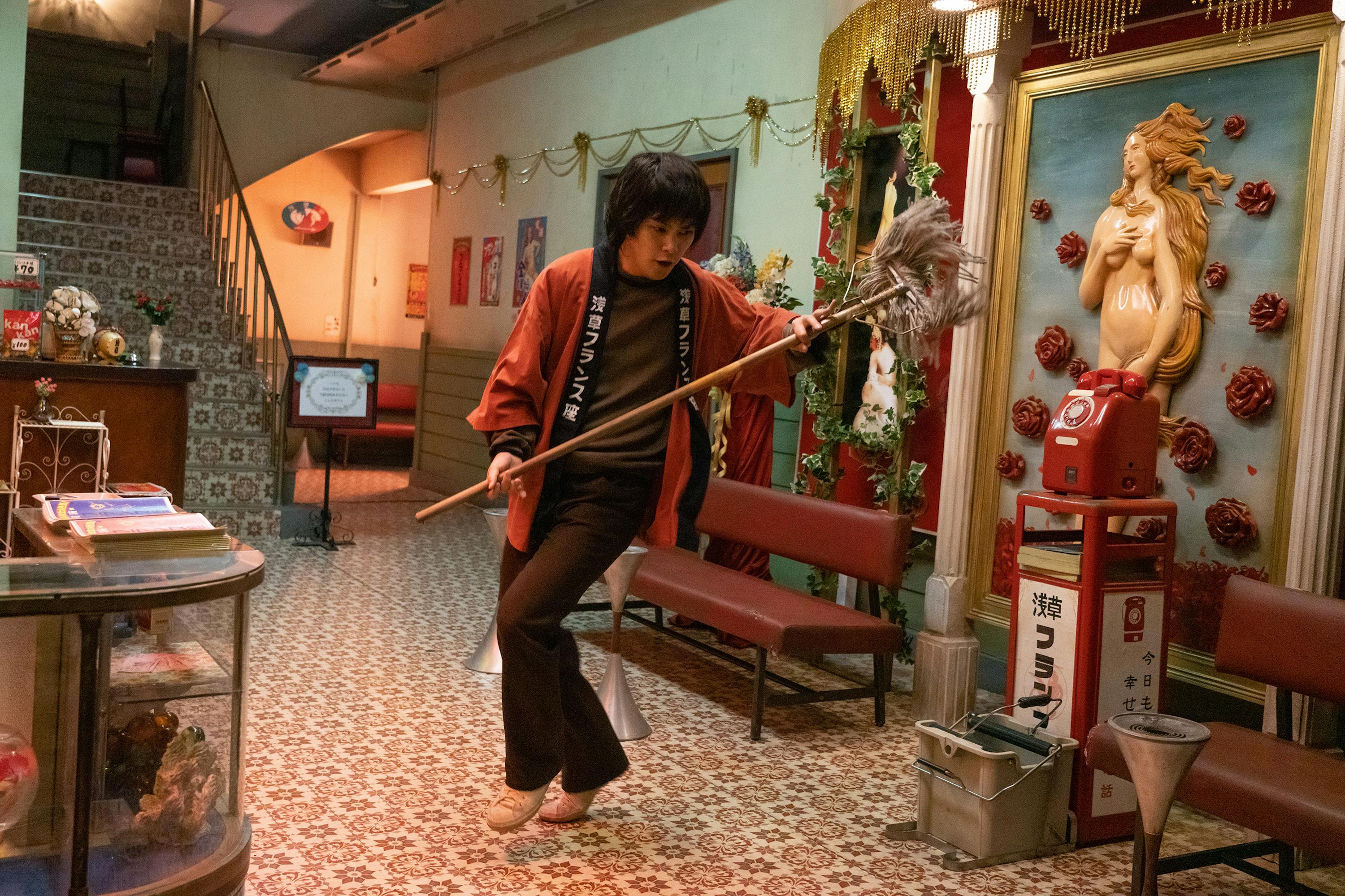 Yuya Yagira wears brown pants and shirt, a red rob, and carries a duster. He dances in the foyer of a house. There is a nude woman on the wall, a phone, and lots of knick-knacks.