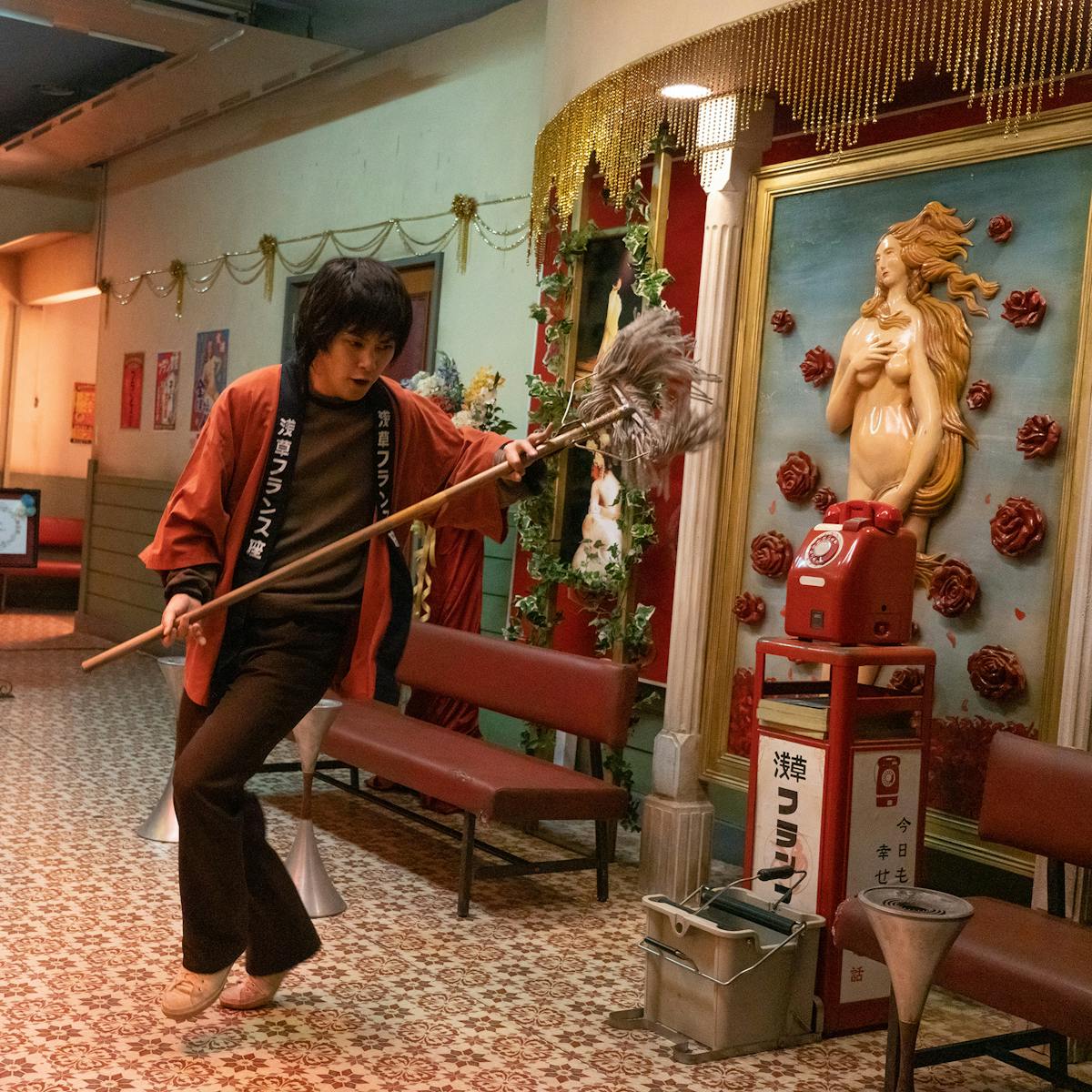 Yuya Yagira wears brown pants and shirt, a red rob, and carries a duster. He dances in the foyer of a house. There is a nude woman on the wall, a phone, and lots of knick-knacks.