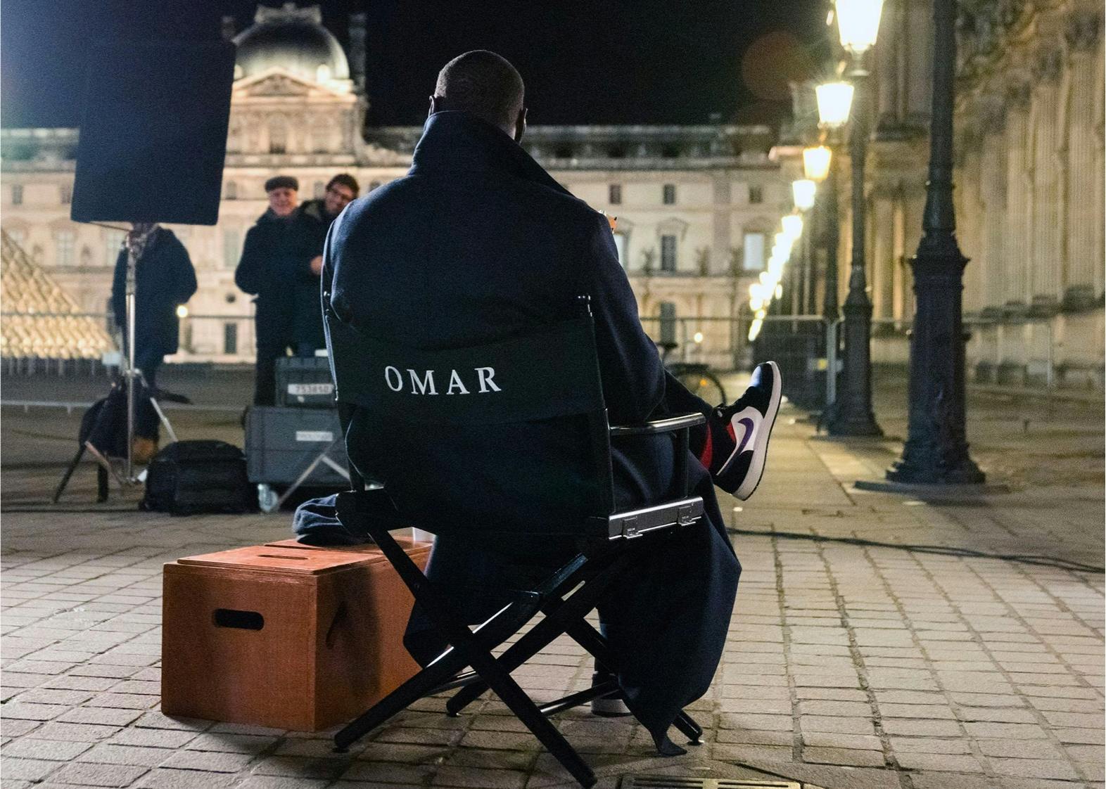 Omar Sy sits in a black set chair with his name embroidered in white on the back. He wears dark clothing and his signature jordans. The nighttime scene is lit by lamps, and the Louvre pyramid is visible to the left. Crew mingle in front of Omar.