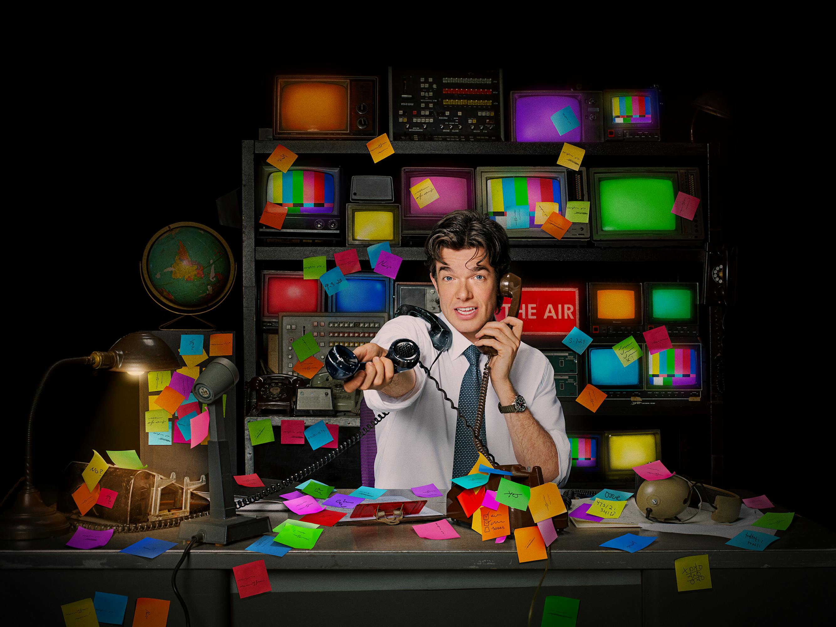 John Mulaney holds a landline out to someone in a post-it covered office.