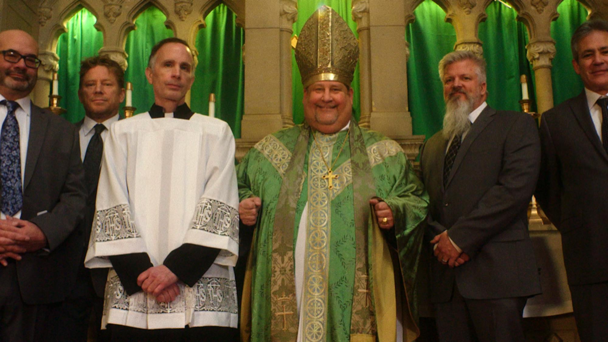 Joe Eldred, Ed Gavagan, Michael Sandridge, Tom Viviano, Dan Laurine, and Mike Forman stand together in a church. Eldred wears a grey suit and thick black glasses, Gavagan wears a dark tie, Sandridge wears a white priest covering and dark clothes underneath, Viviano wears green robes and a bishop hat, Laurine wears a dark suit and has a grey beard.
