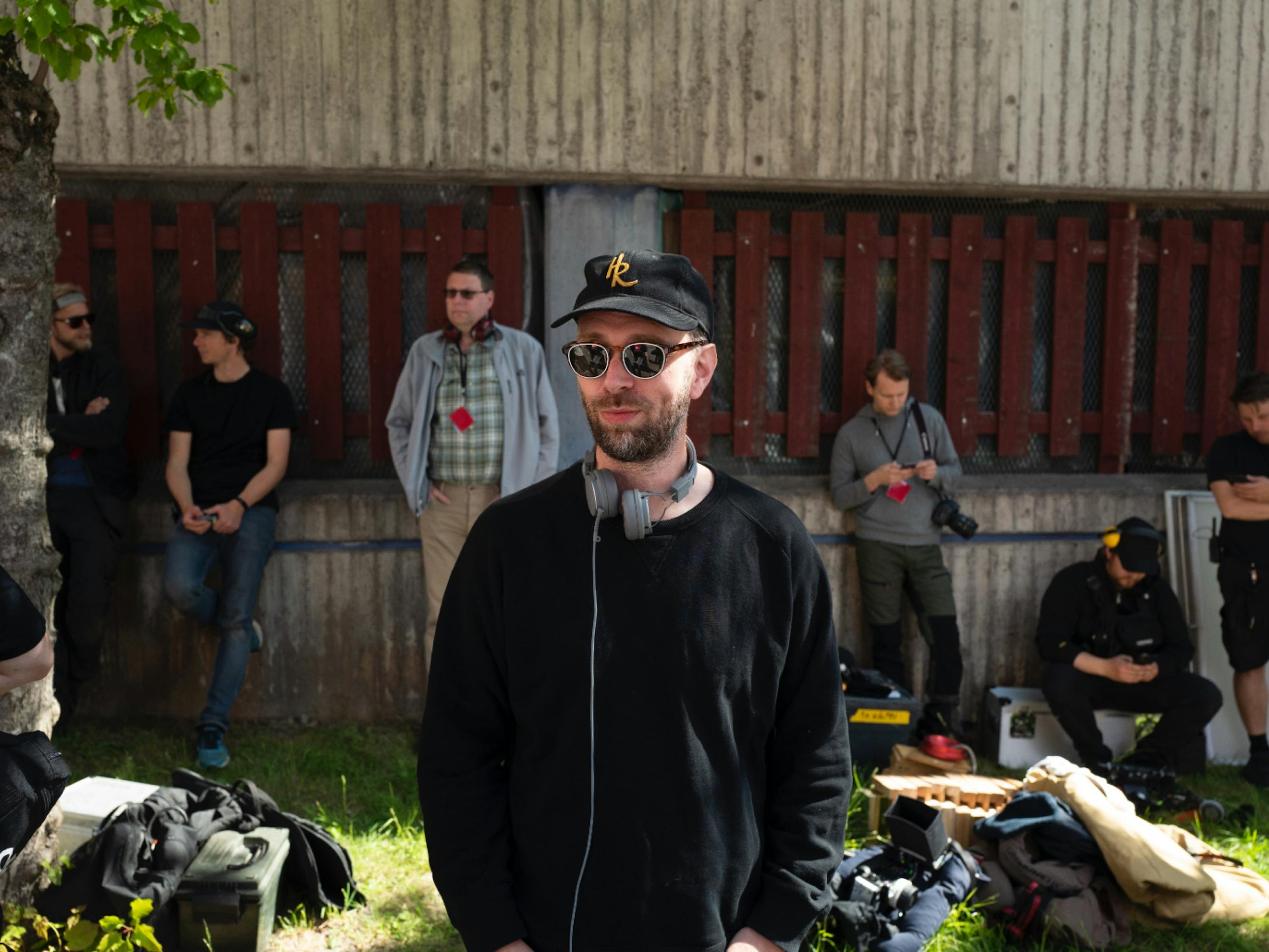 Co-creator and director Jesper Ganslandt on the set of Snabba Cash. He wears a black longsleeve, black hat, sunglasses, and earphones around his neck. There are seven other crew people in the background equipped with cameras and lanyards.