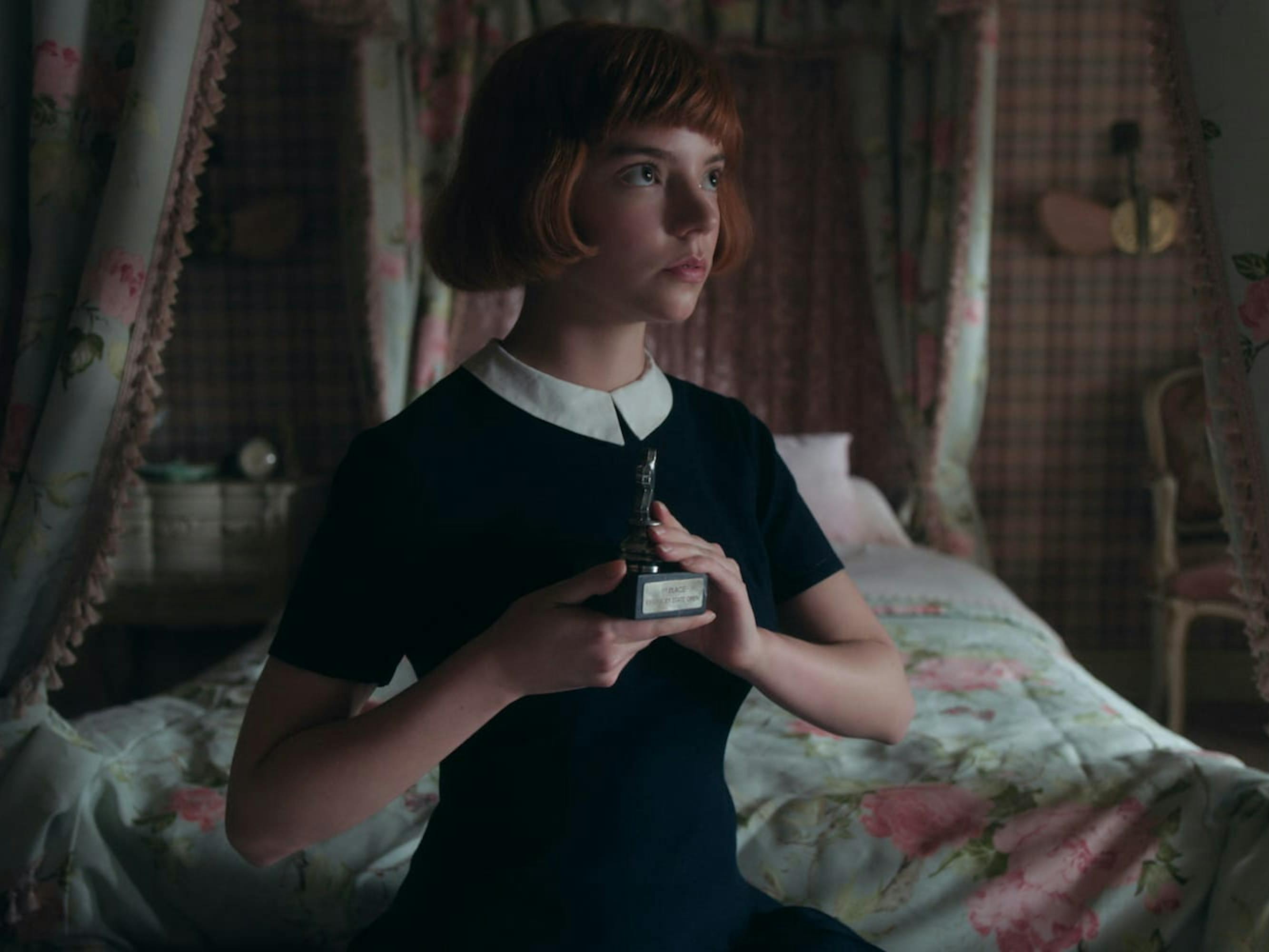 Beth Harmon (Anya Taylor-Joy) sits in her floral and plaid decorated bedroom and holds a small trophy to her chest. She wears a navy dress with a white color and sits erect.