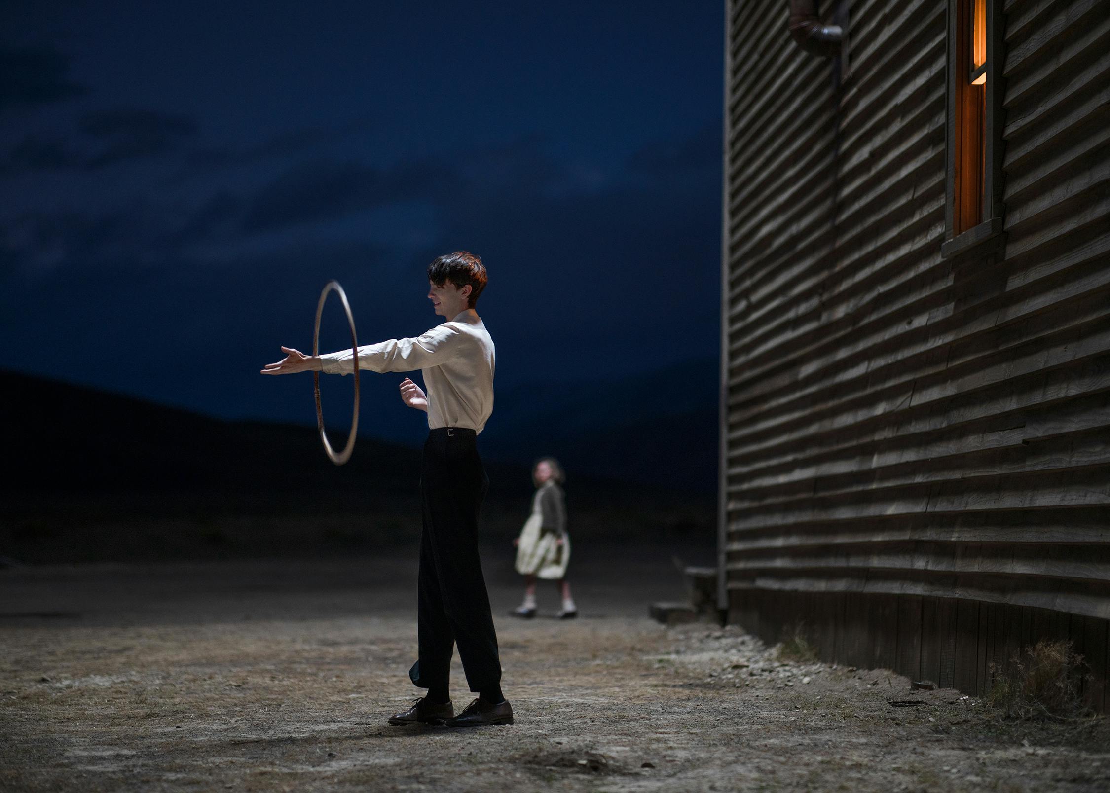 Kodi Smit-McPhee stands outside in the dark, wearing black pants and a white shirt. He balances a hula hoop on his arm, as a woman walks by him in the distance.