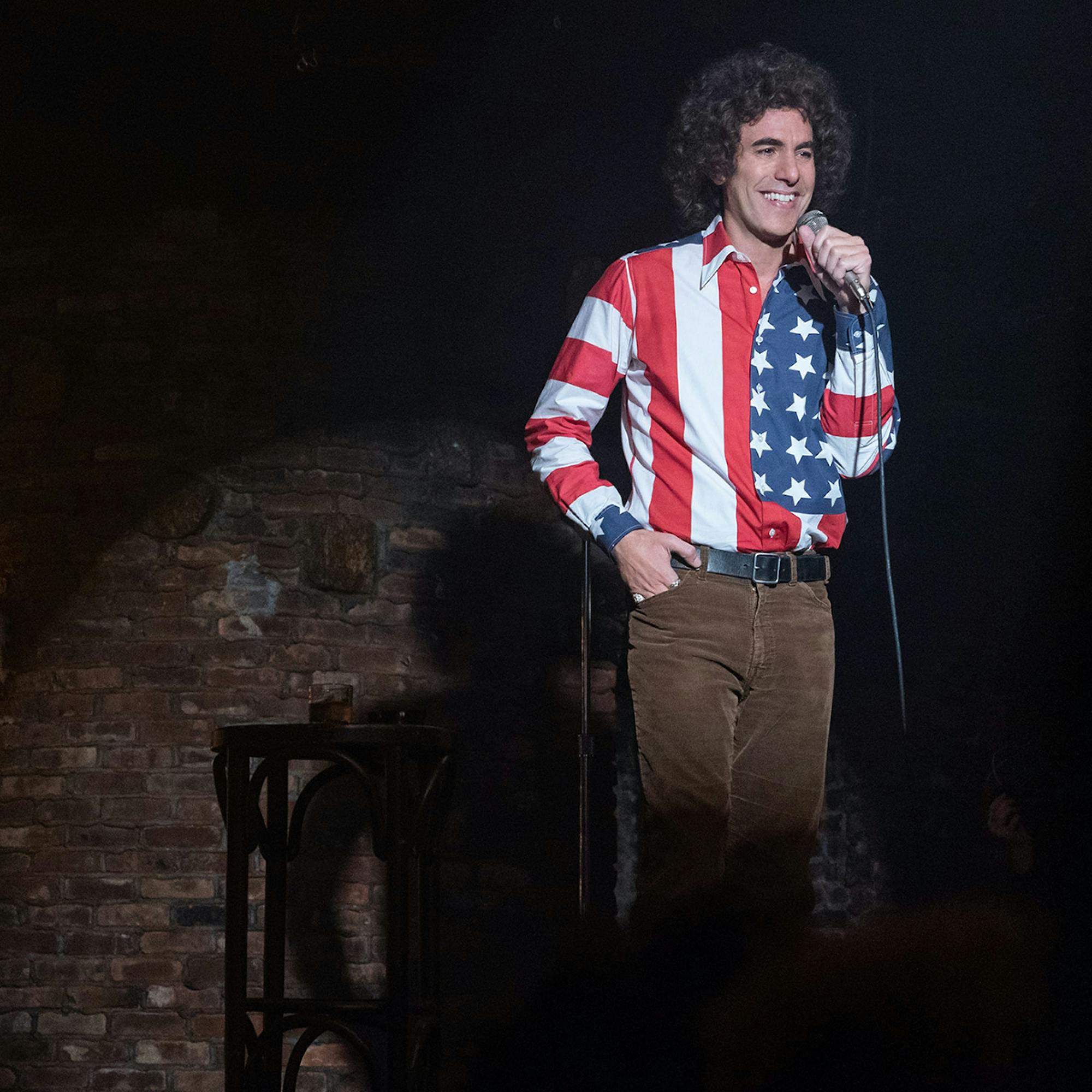 Cohen wears an American flag shirt and brown cords, and stands behind a microphone. His hand is in his pocket, and he is laughing. There is a single spotlight lighting him up but everything else is in the shot is dark. 