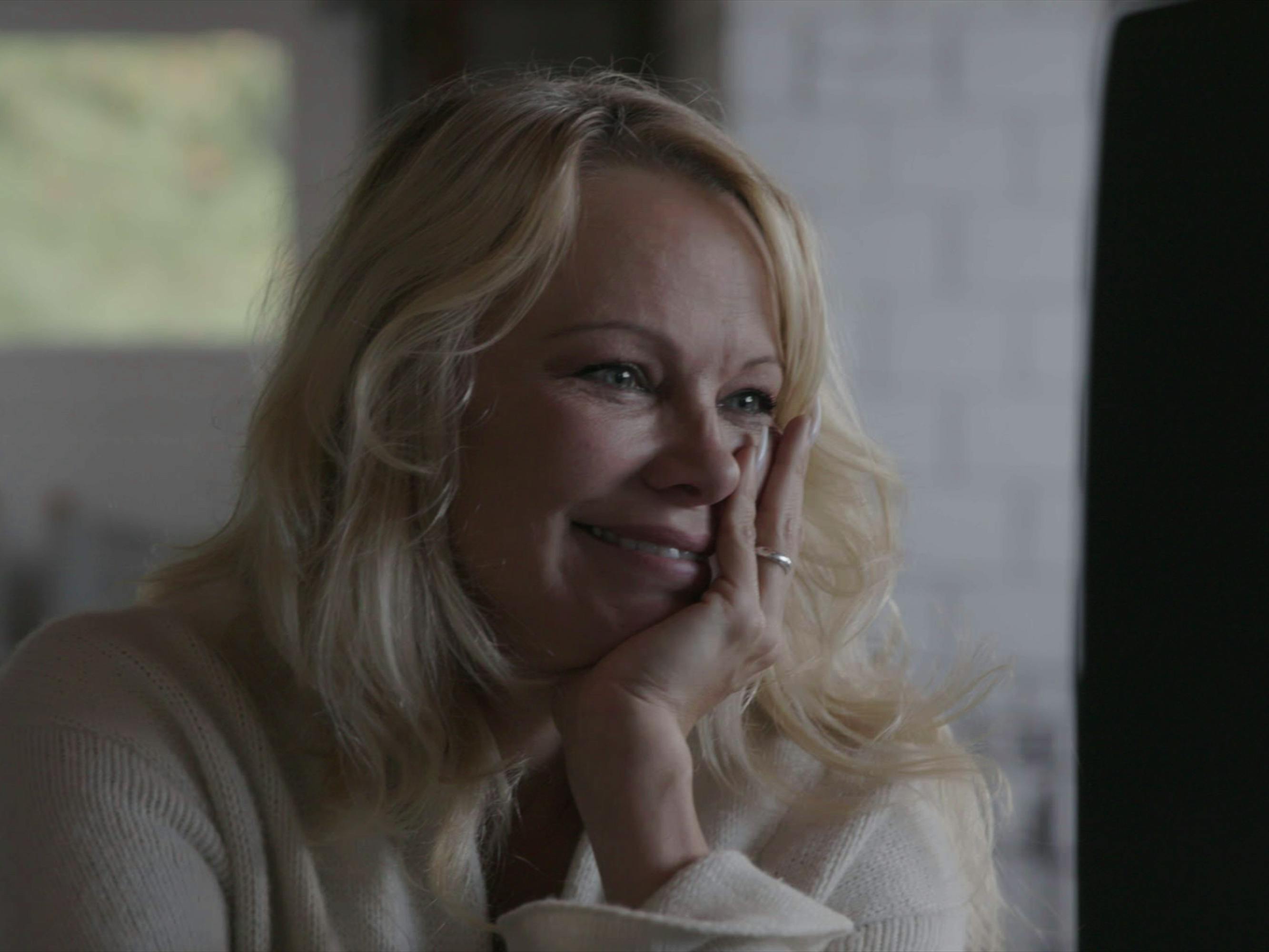 Pamela Anderson looks quietly beautiful in this cozy, morning-lit kitchen shot. She holds her hand to her face and smiles at something off-camera.