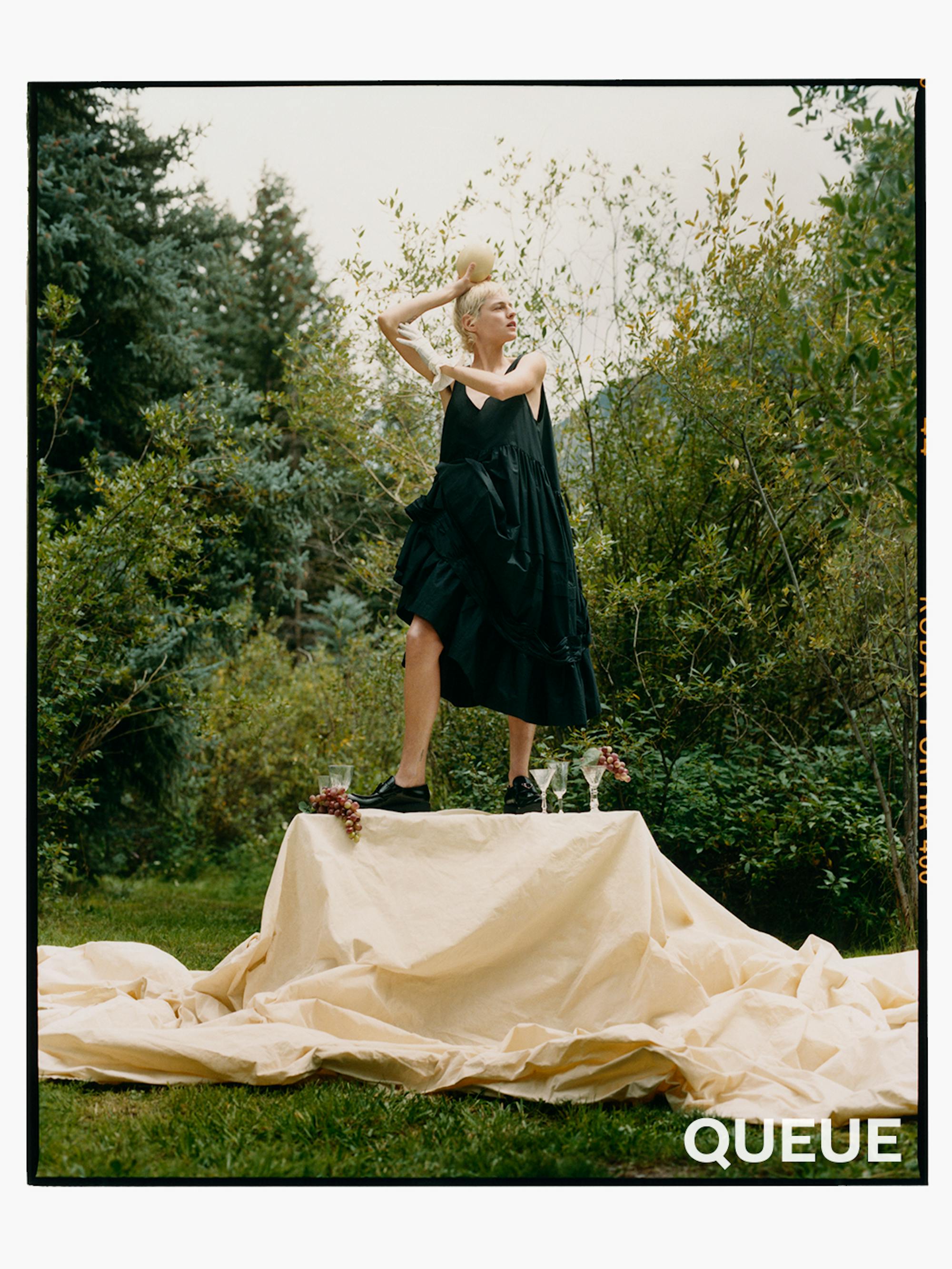 Emma Corrin wears a black dress and stands on a raised platform in the woods. 