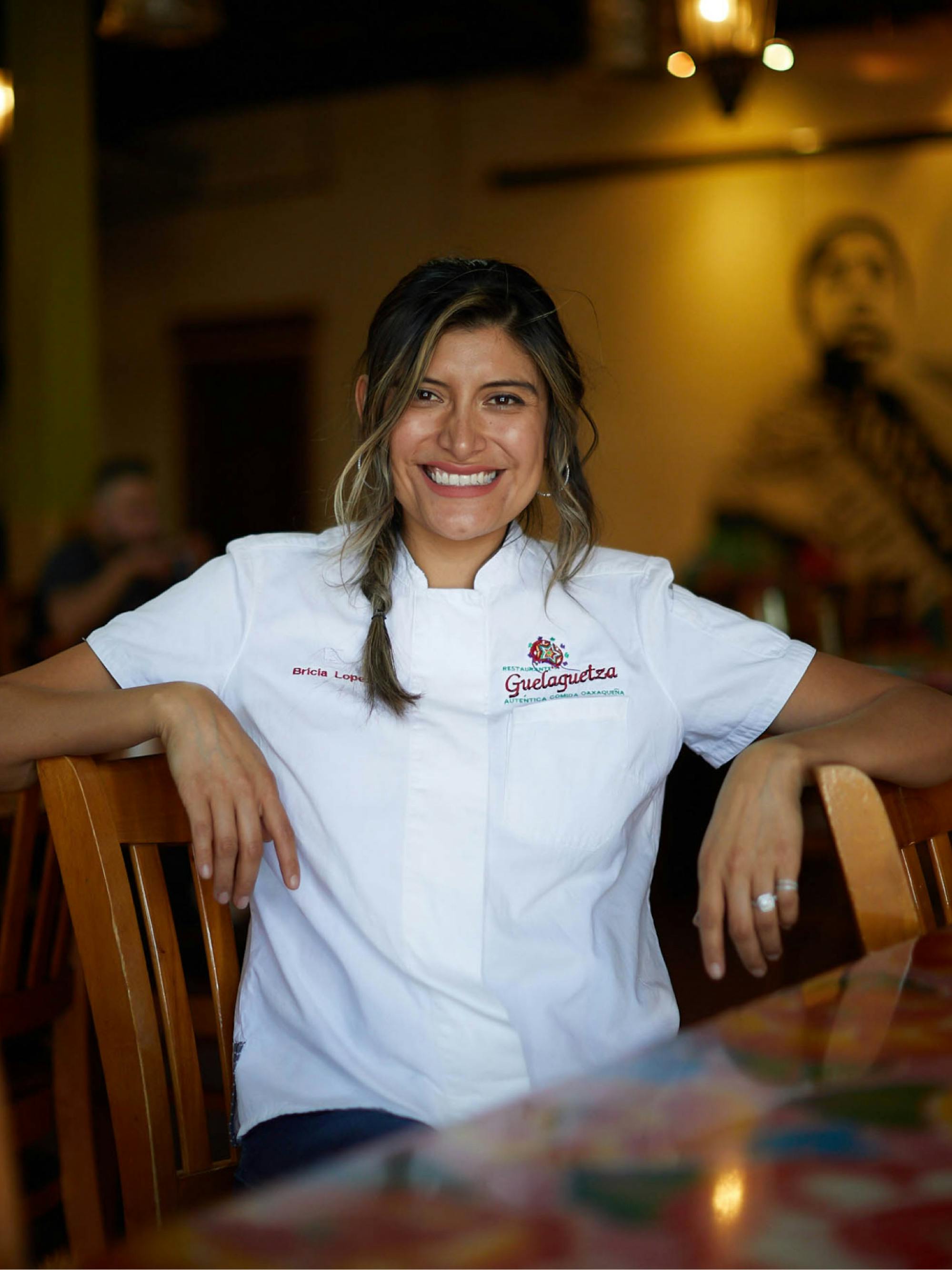 Lopez at her Los Angeles Restaurant, Guelaguetza. She’s smiling and sitting at one of the tables, with her arms propped up on two chairs. She wears a white chef’s shirt with the restaurant’s logo. There’s a mural on a yellow wall in the background.