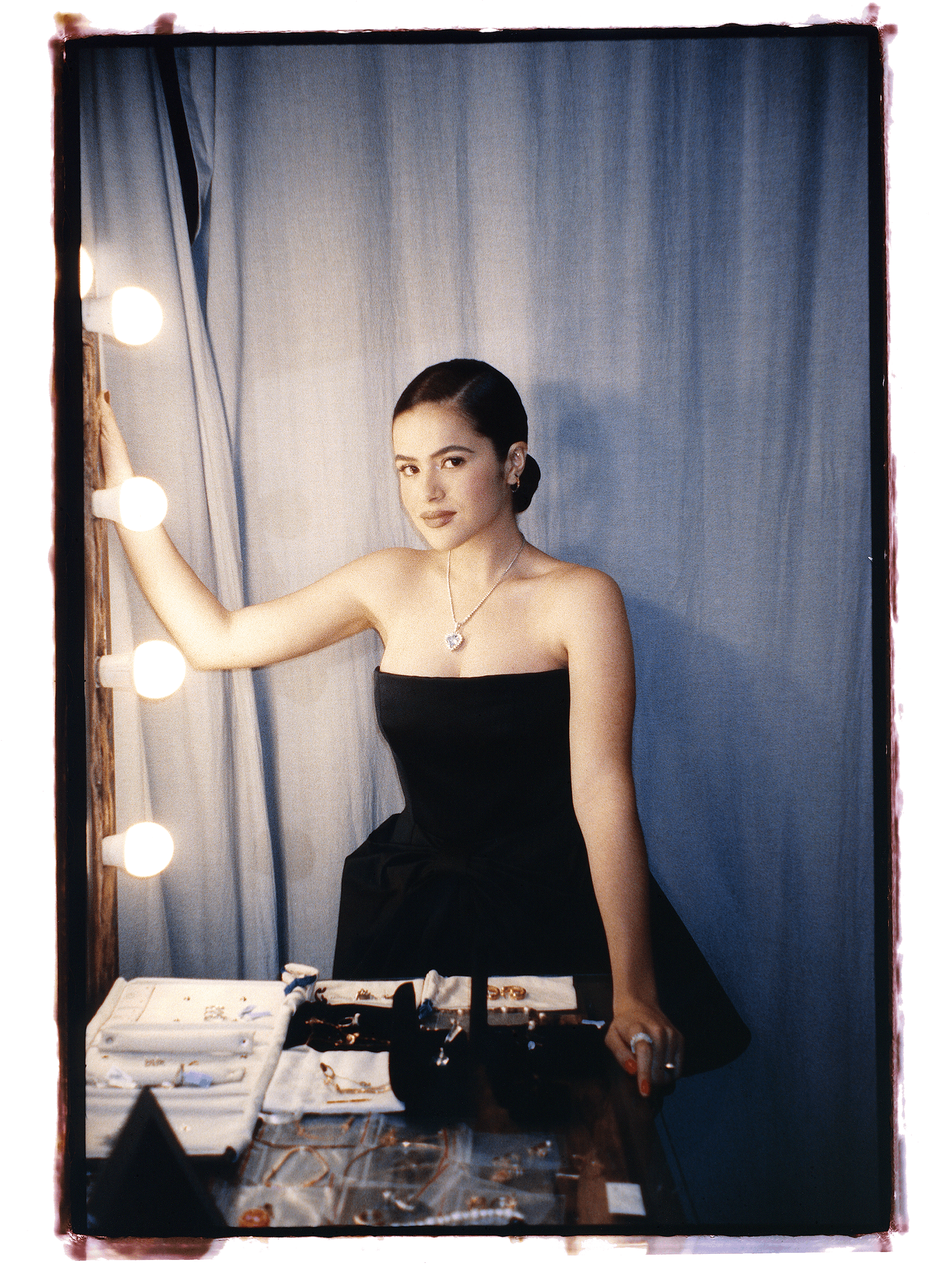 Maisa Silva wears a black strapless dress and stands by a bulb-lit mirror. She stands against a blue curtain.