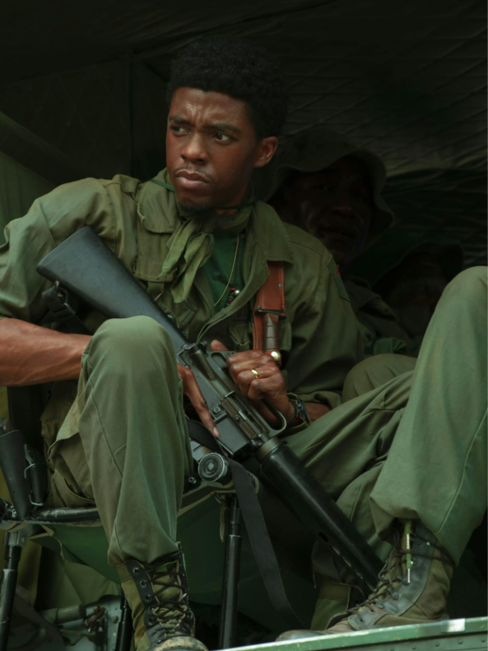 Boseman as Stormin’ Norman, wearing combat gear, boots, and holding a very large gun. He sits in a green truck and looks at something off camera. 