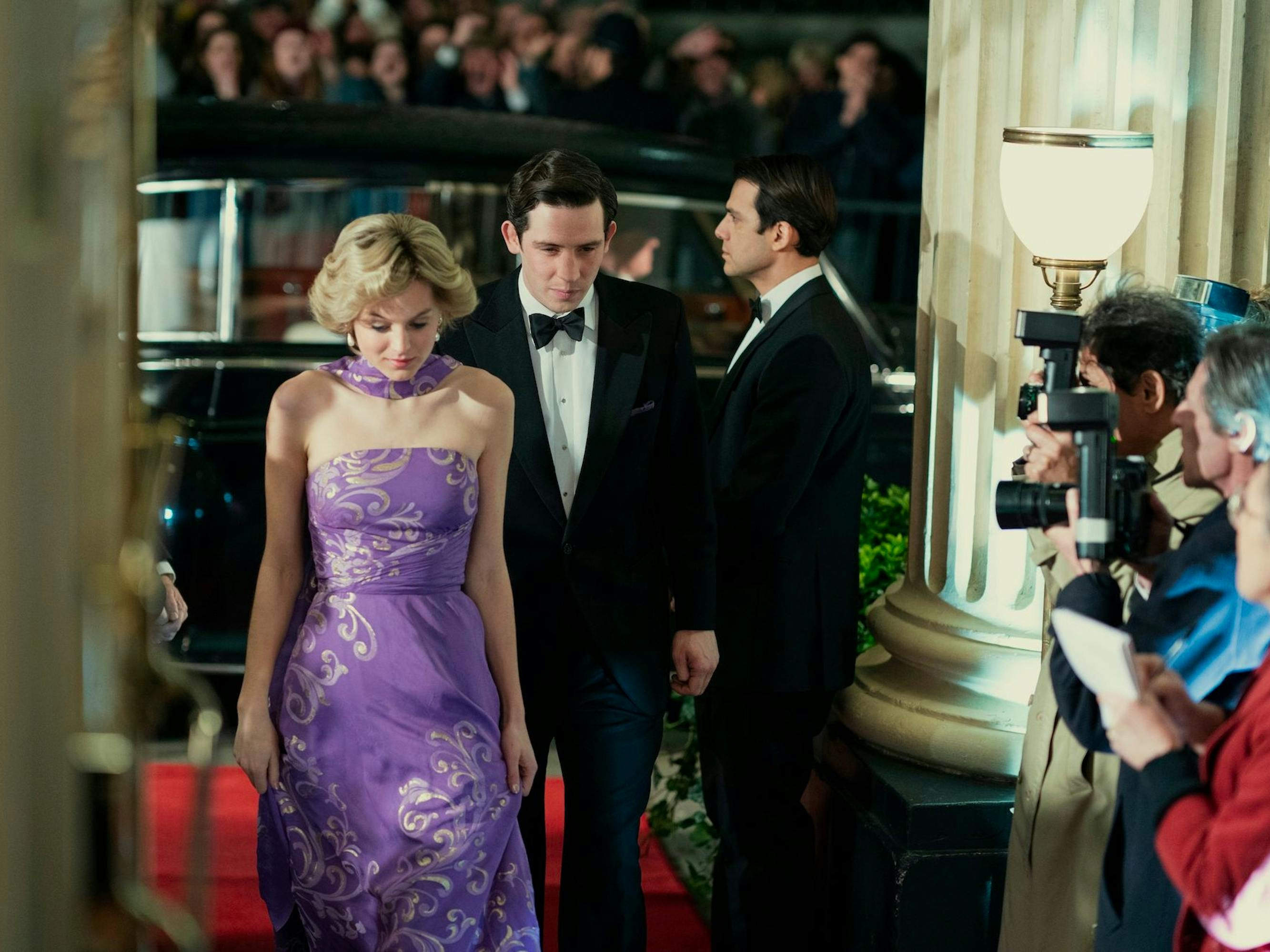 Diana Spencer (Emma Corrin) and Prince Charles (Josh O’Connor) enter through wide white doors. Corrin wears a beautiful purple dress, and Charles wears a tux. Outside are hundreds of fans and reporters, and inside are more cameras poised to snap a picture.