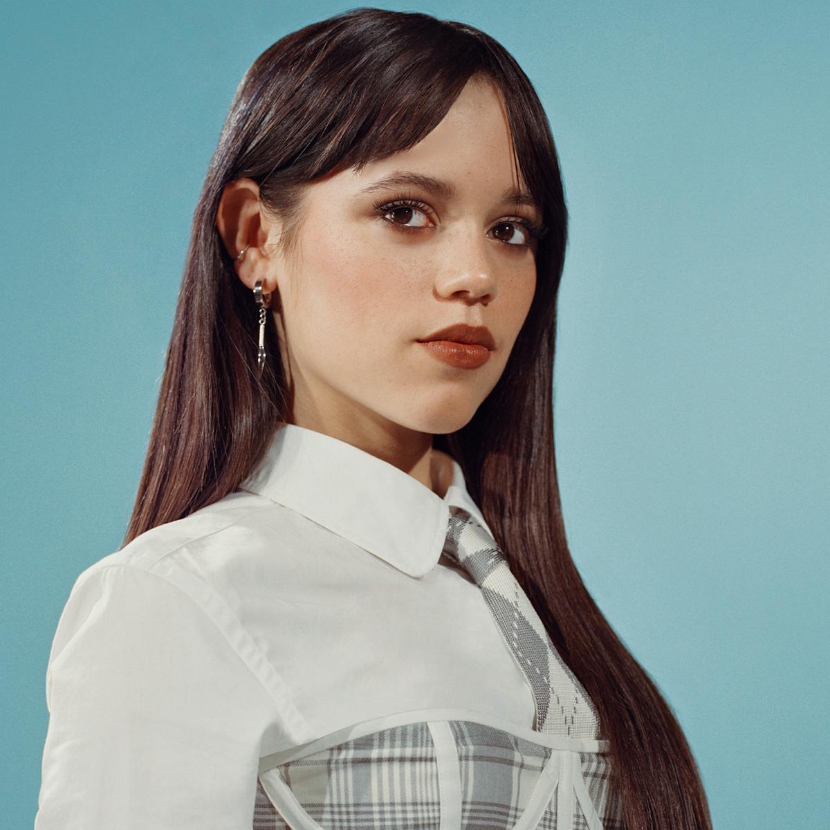 Jenna Ortega wears a white shirt with a gray-and-white plaid dress over it. She stands against a teal background.