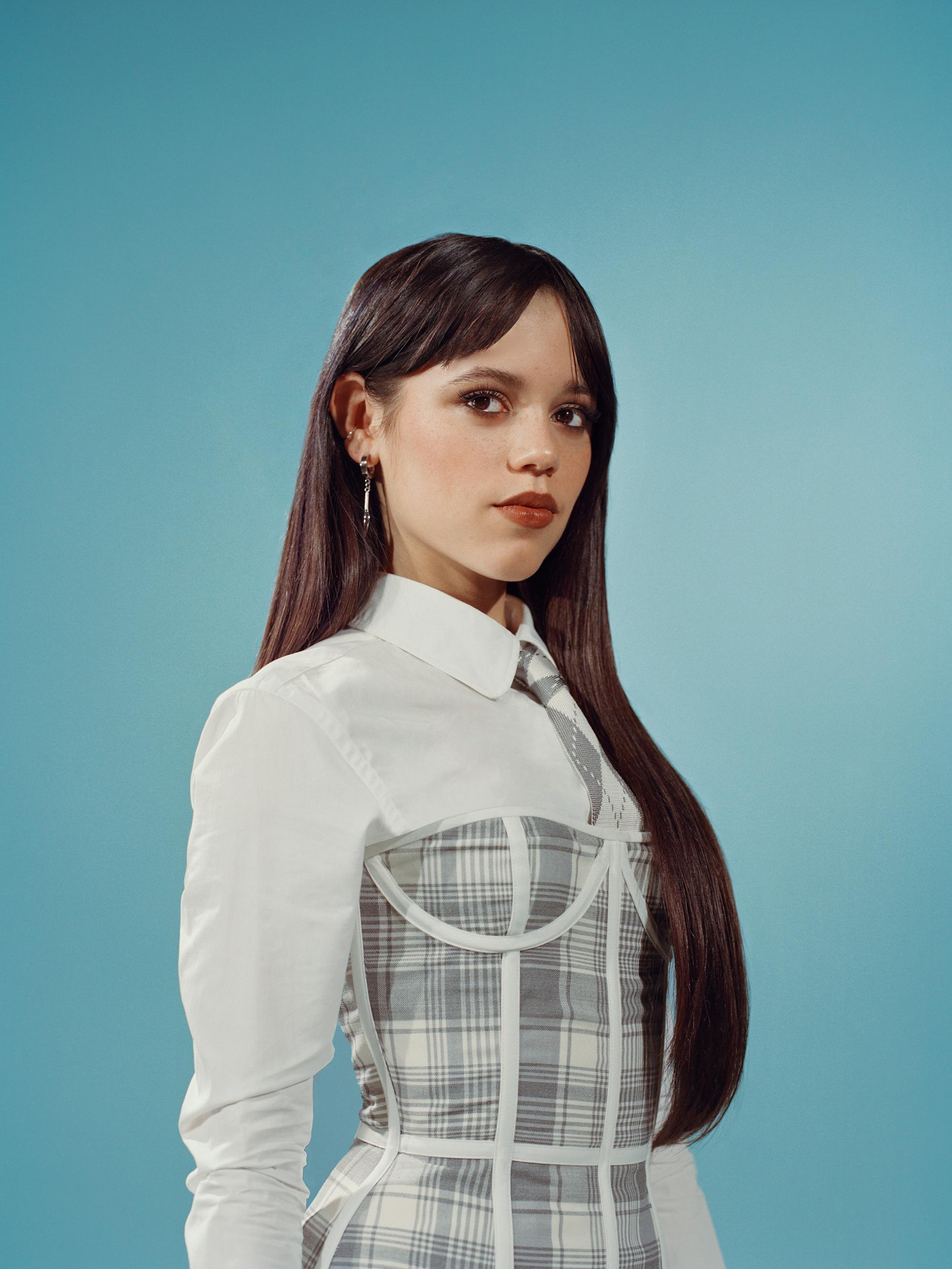 Jenna Ortega wears a white shirt with a gray-and-white plaid dress over it. She stands against a teal background.