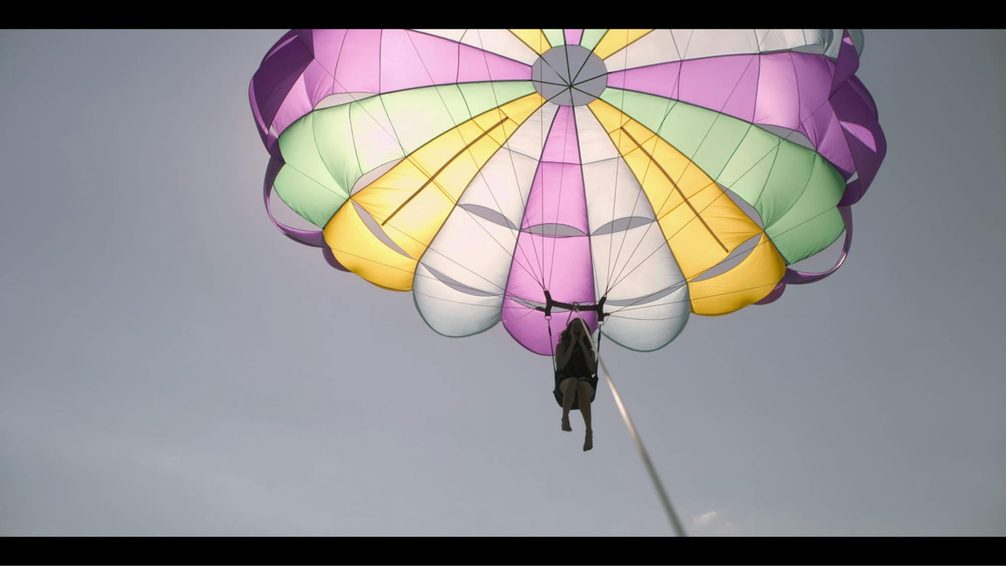 Sara (Ximena Lamadrid) parasails in a clear blue sky. Her contraption is purple, white, yellow, and green. Her figure is shadowy but you can sense her excitement from her hands framing her face.