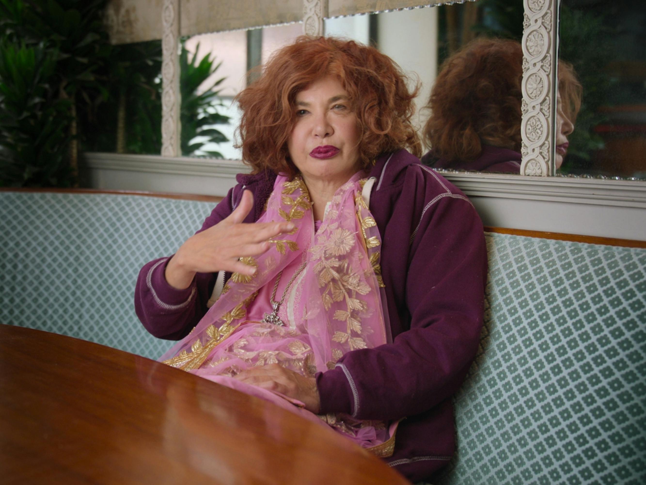 Tama Janowitz sits in a booth wearing a purple sweater and pink scarf.