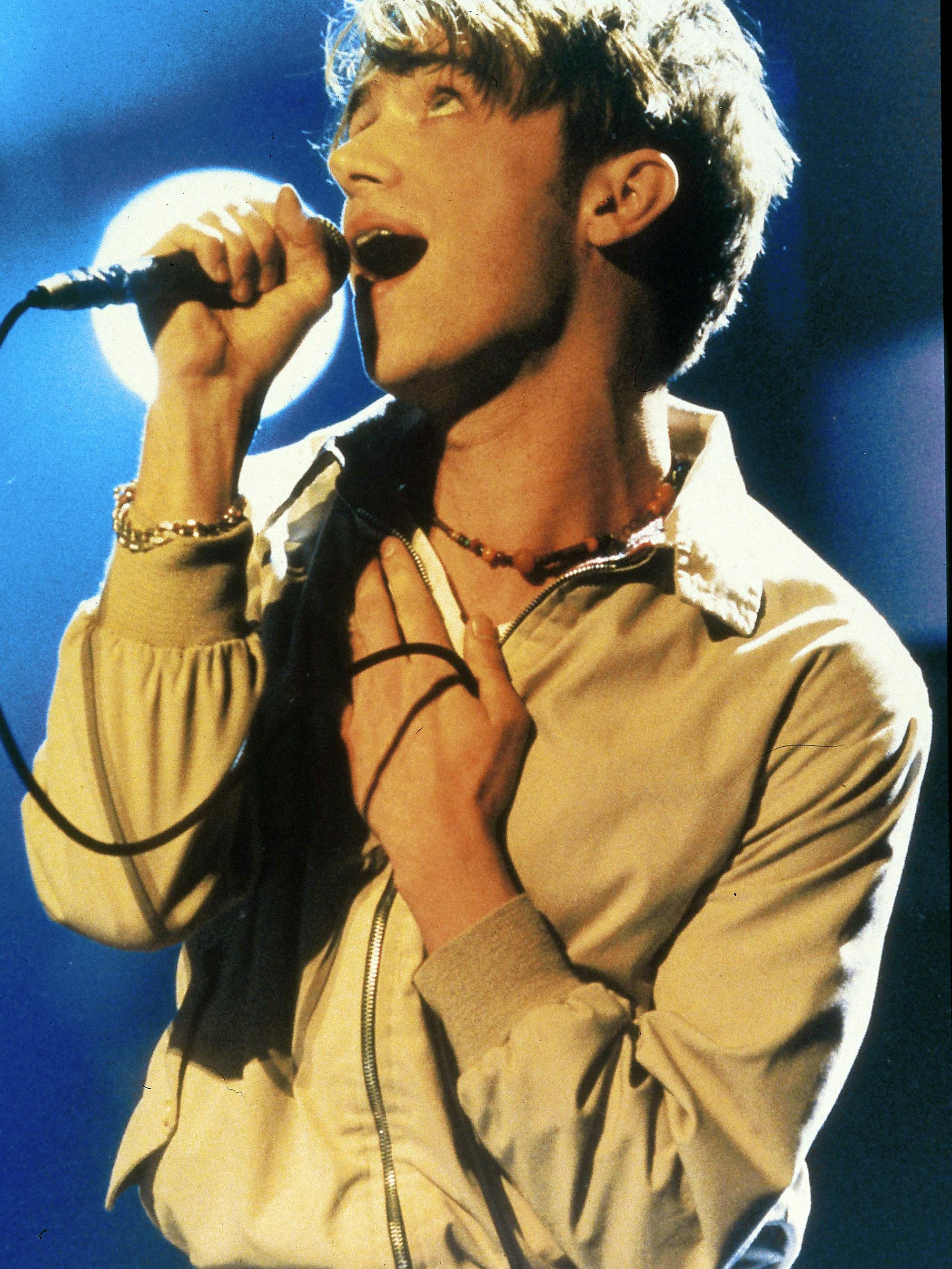 Damon Albarn sings into a microphone with his hand on his chest. 